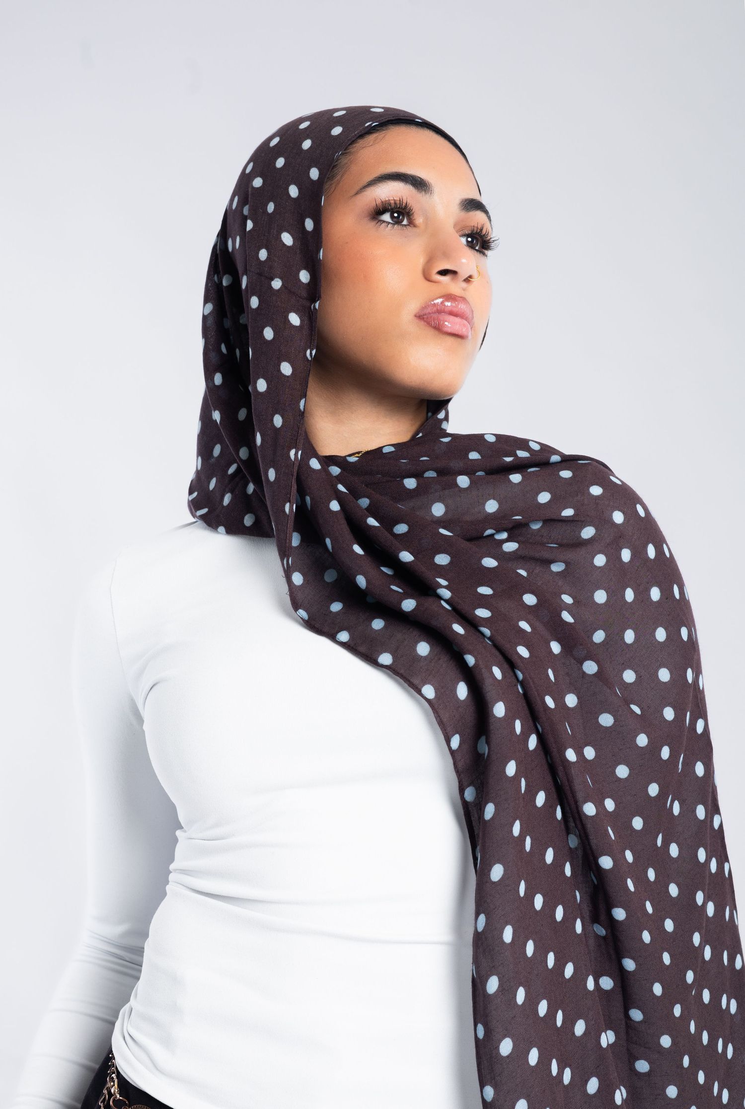Woman wearing a dark brown polka dot hijab with light blue spots paired with a white long-sleeve top.