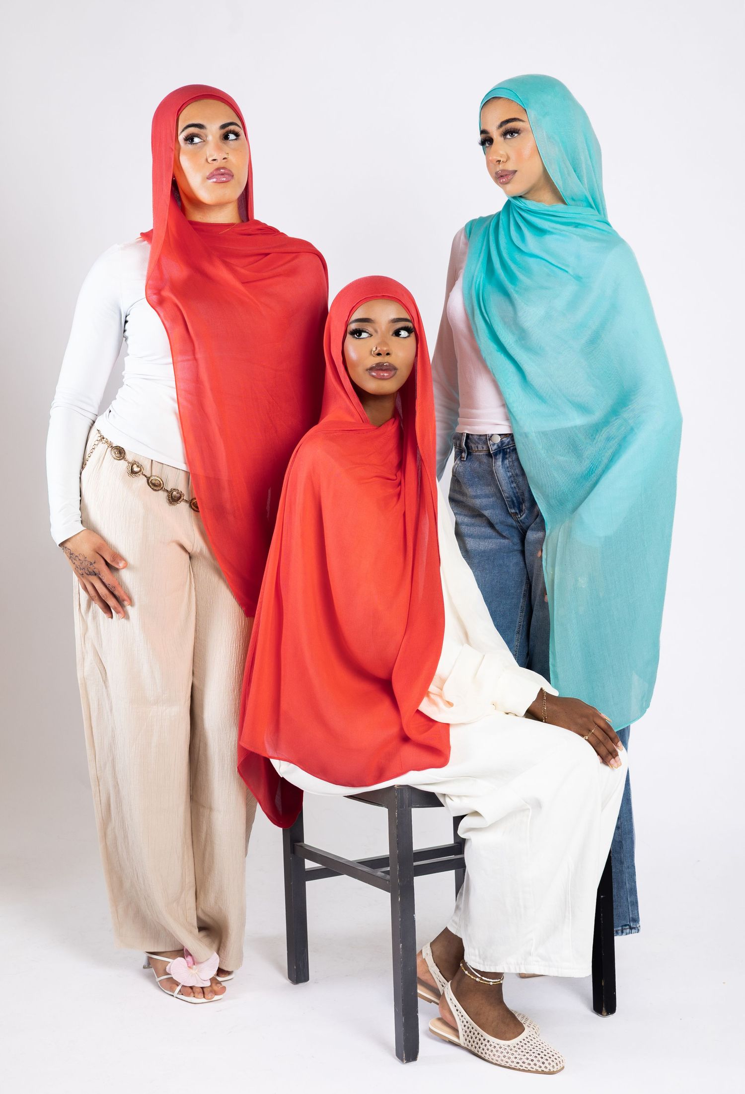 Three women wearing colorful hijabs in red, coral, and teal pose elegantly in a studio setting.