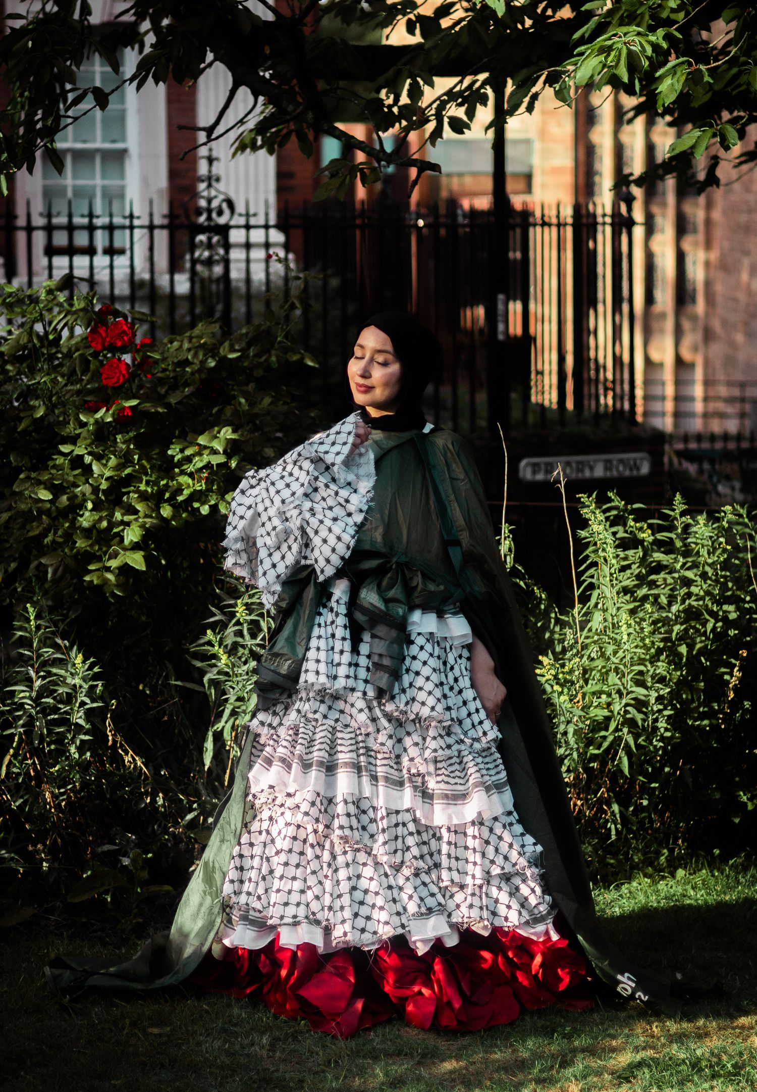 Victorian-style dress with checkered pattern, green shawl and red trim photographed in a garden setting with ornate iron fence.