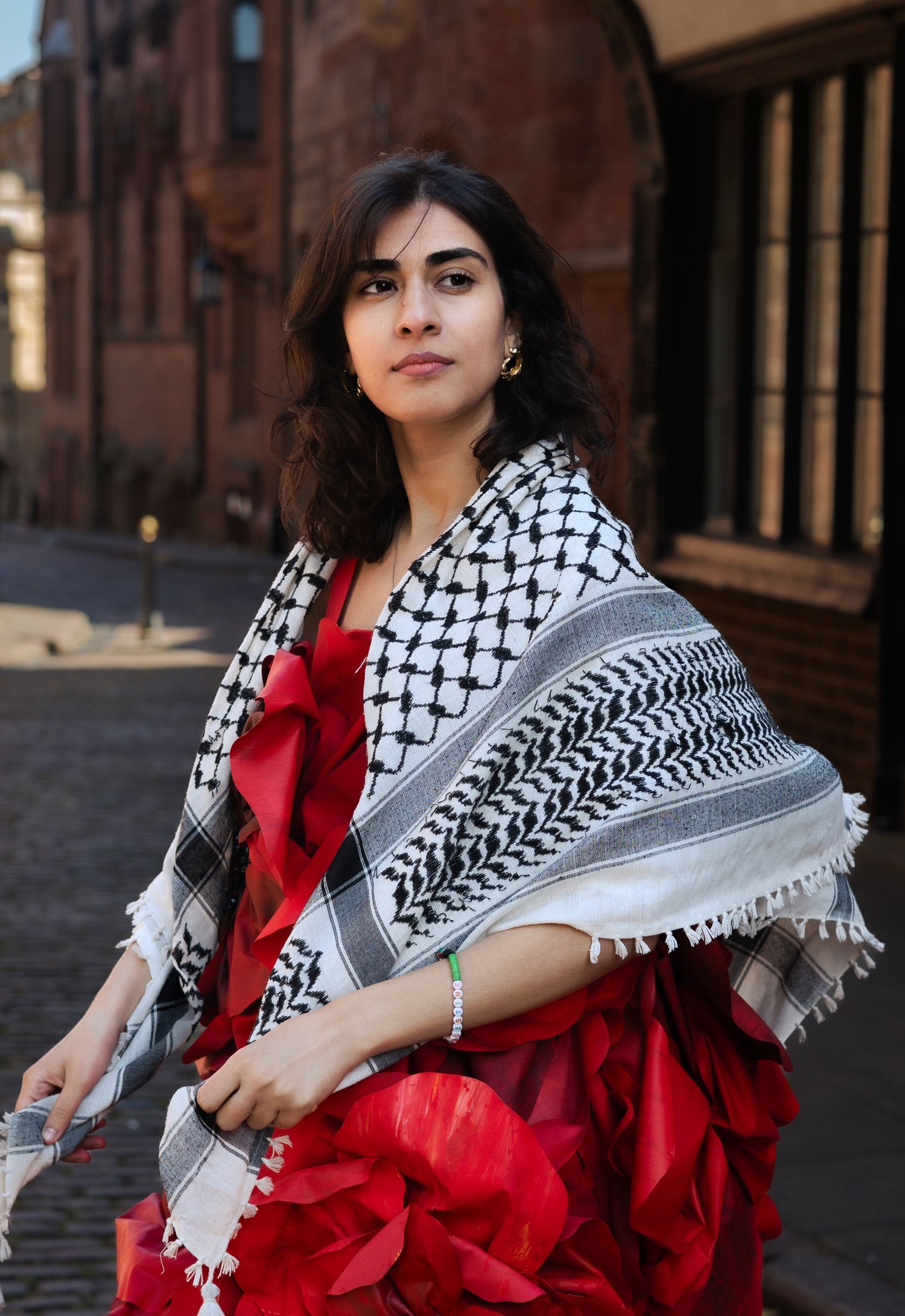 Model draped in traditional patterned shawl over red formal dress poses in historic setting.