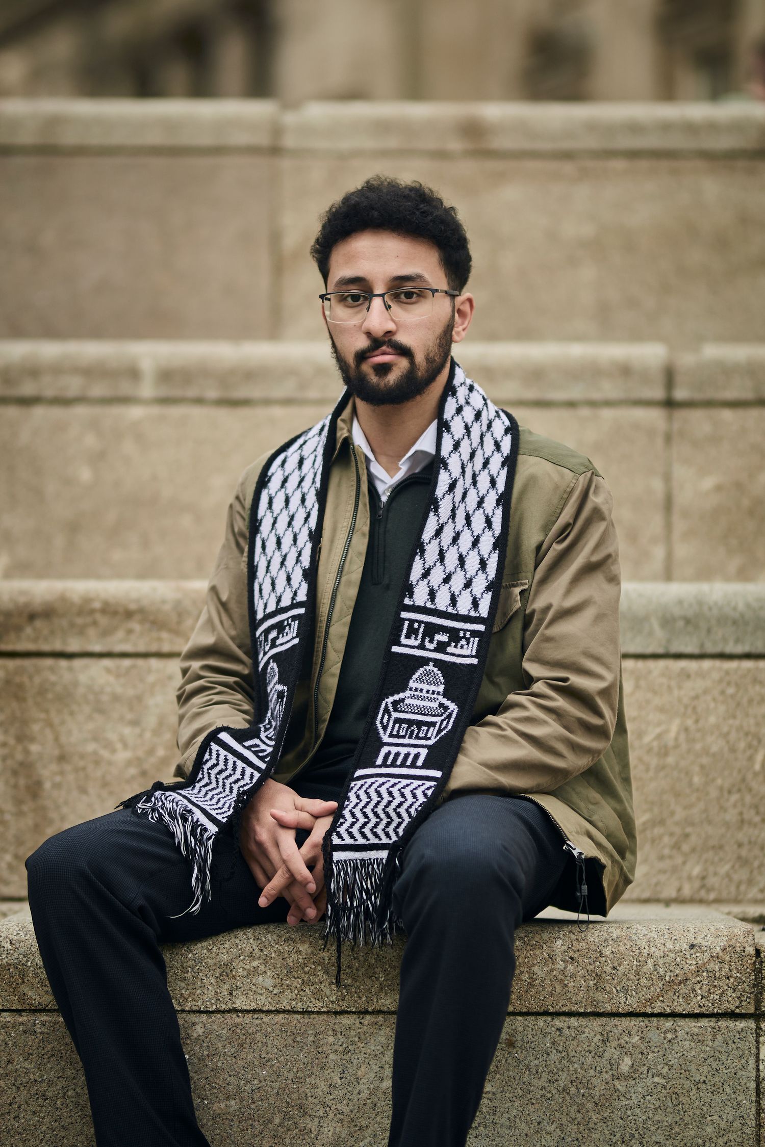 Person wearing keffiyeh scarf sits contemplatively on stone steps.
