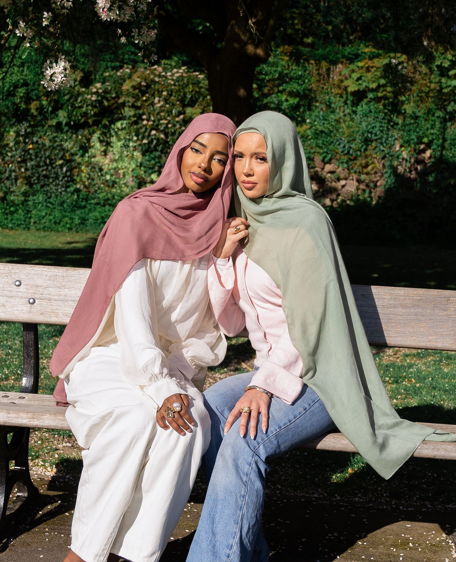 Two women wearing colorful hijabs sit together on a park bench, smiling, dressed in white and blue jeans.