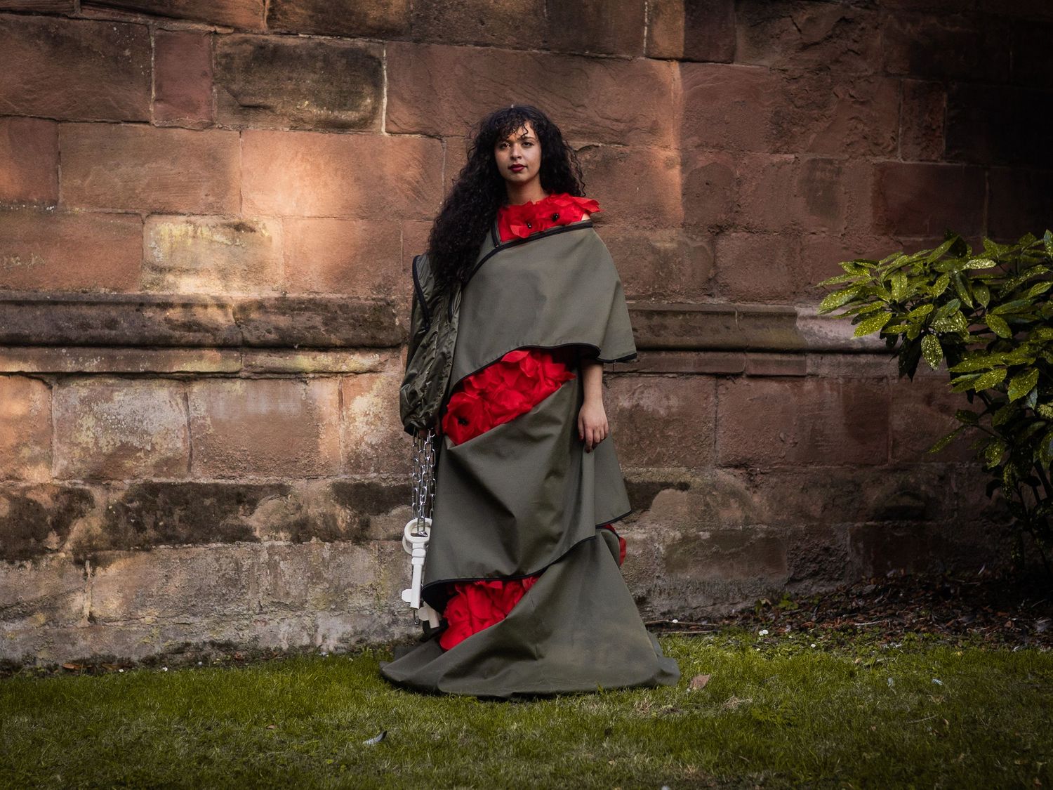 Woman in dramatic olive green layered cape with red floral accents stands before a rustic stone wall.