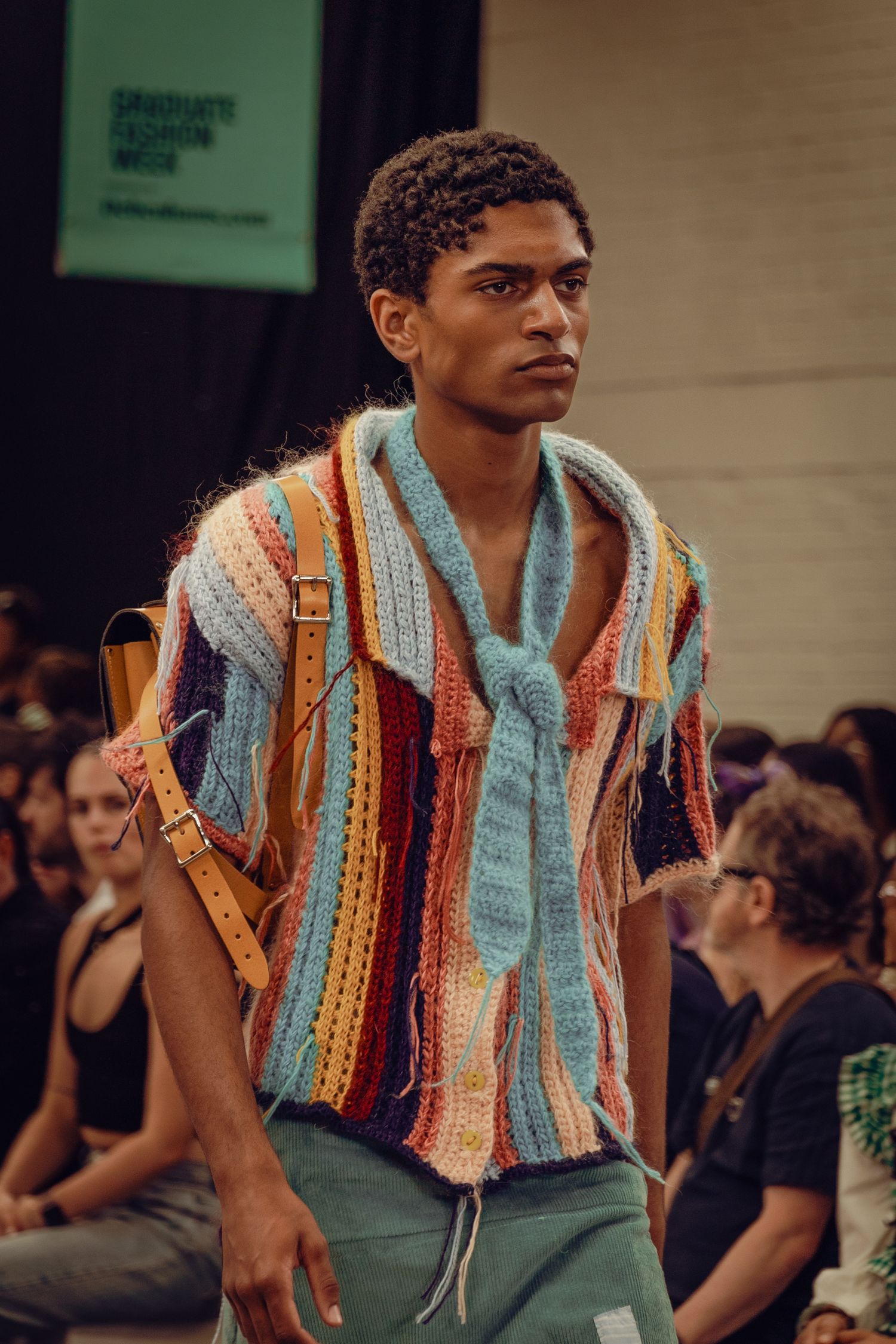 Model wearing colorful striped knit sweater with turquoise scarf on fashion runway.