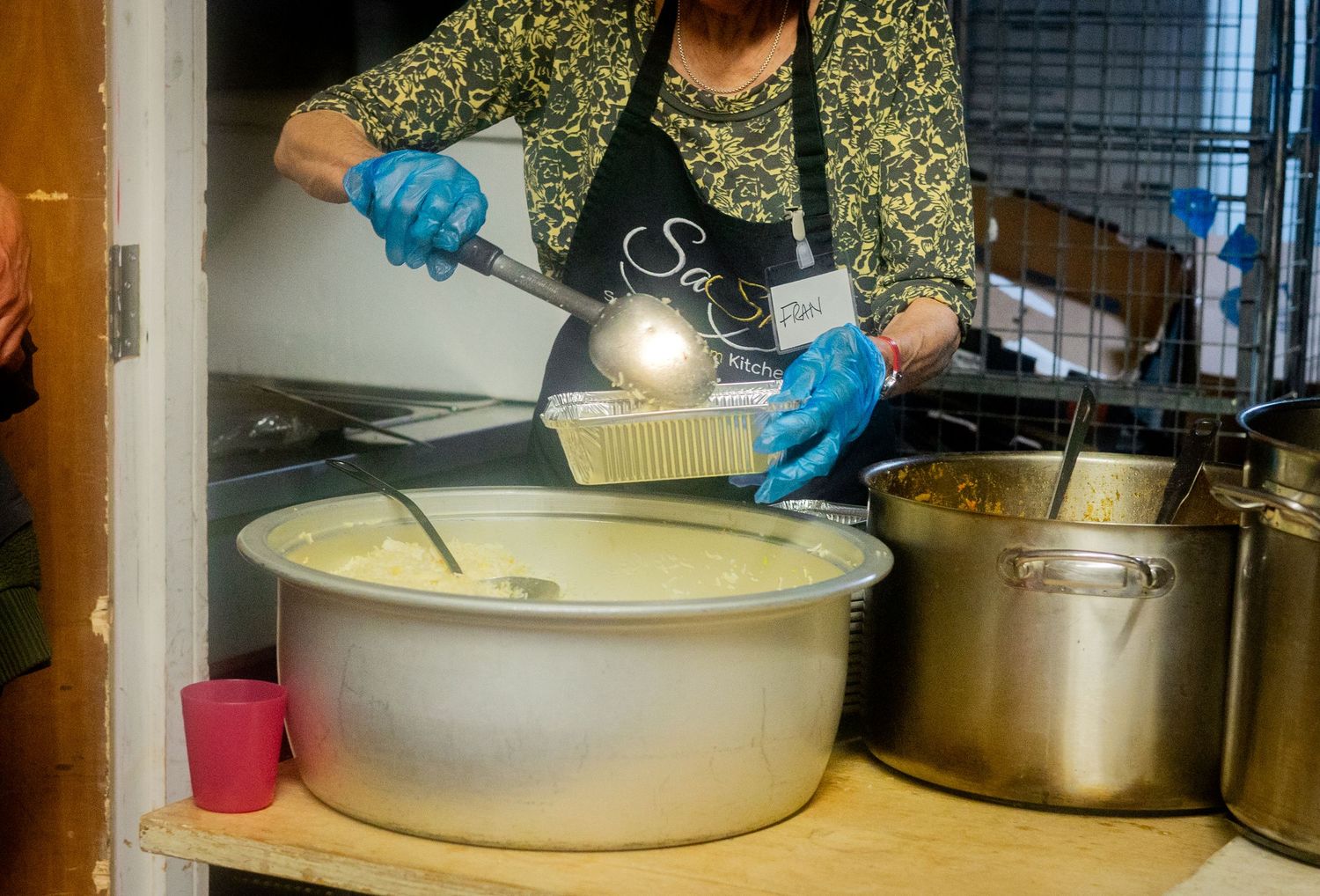 Someone in blue gloves and an apron ladles soup from a large commercial pot in a kitchen.