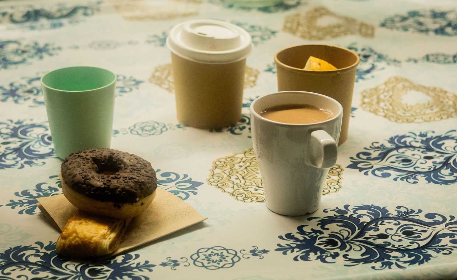 Coffee cups and beverages arranged on a decorative tablecloth at a social gathering.