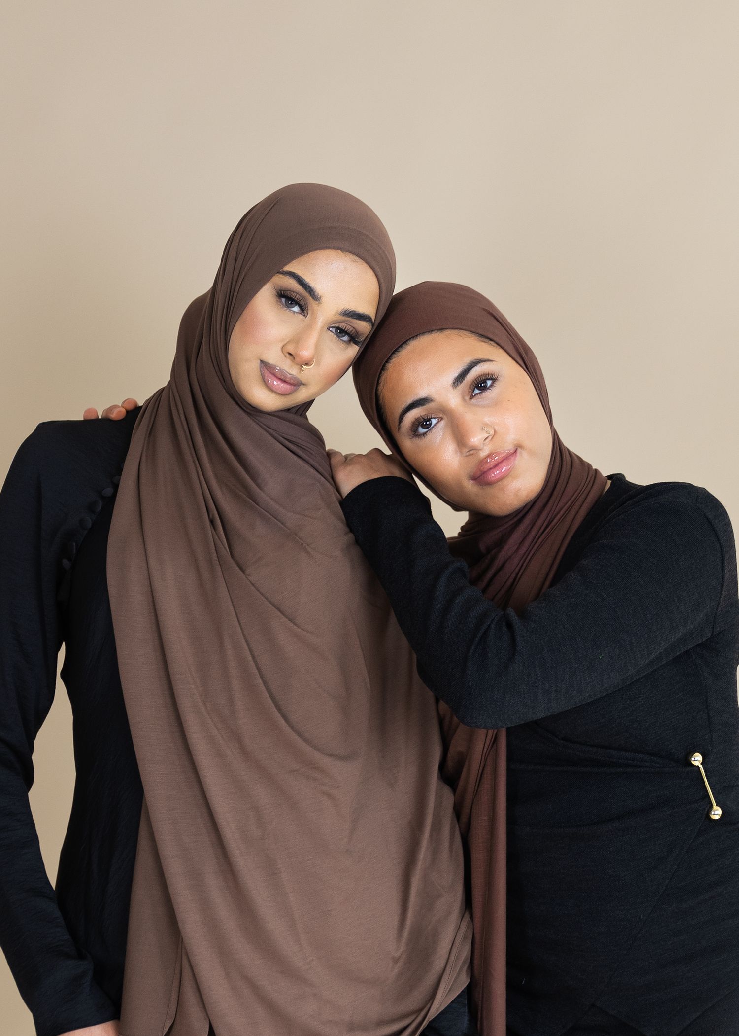 Two people wearing brown hijabs and black sweaters pose together against a neutral background.