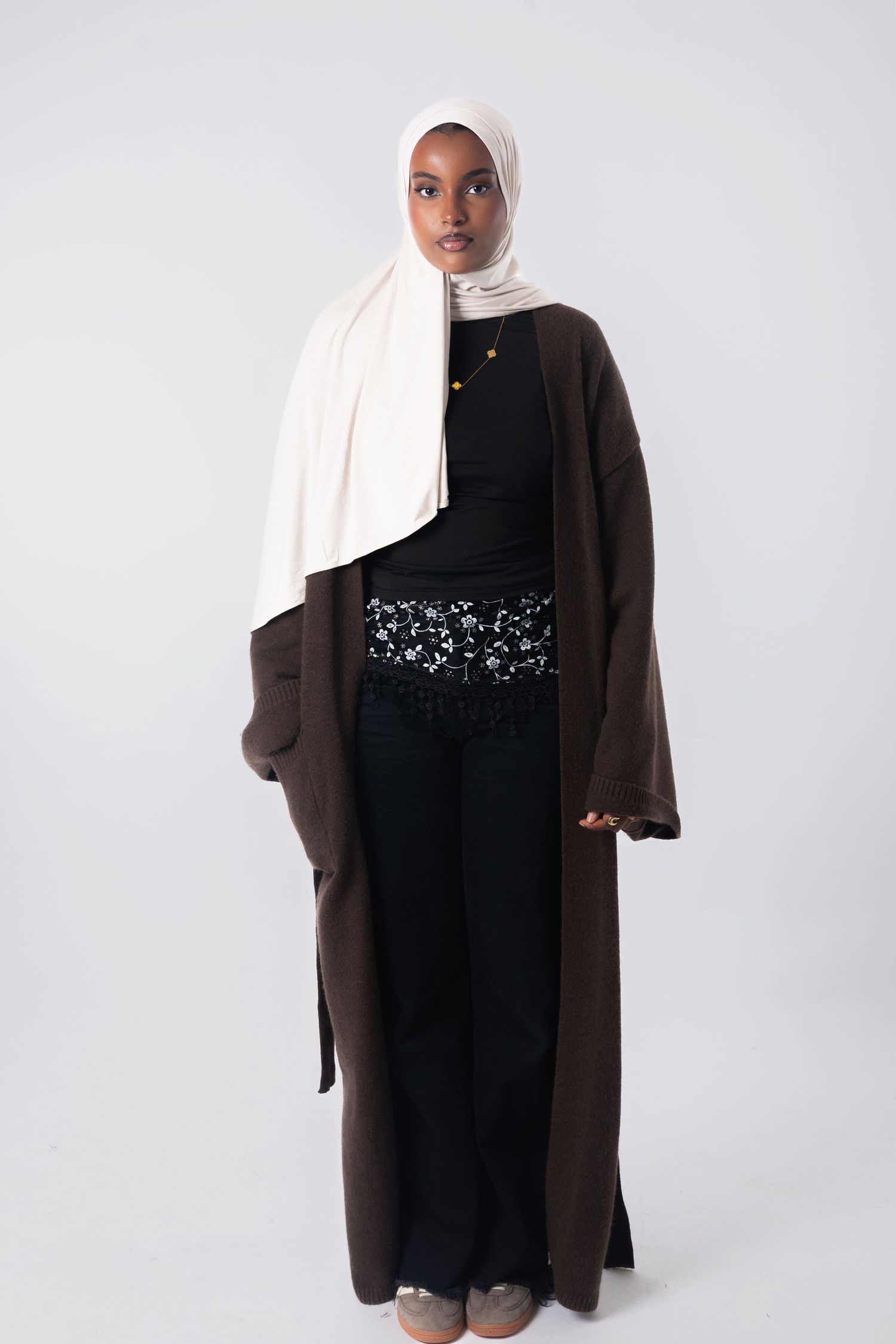 Woman wearing cream hijab, dark brown longline cardigan, black top, lace waistband pants, and gray shoes on light background.