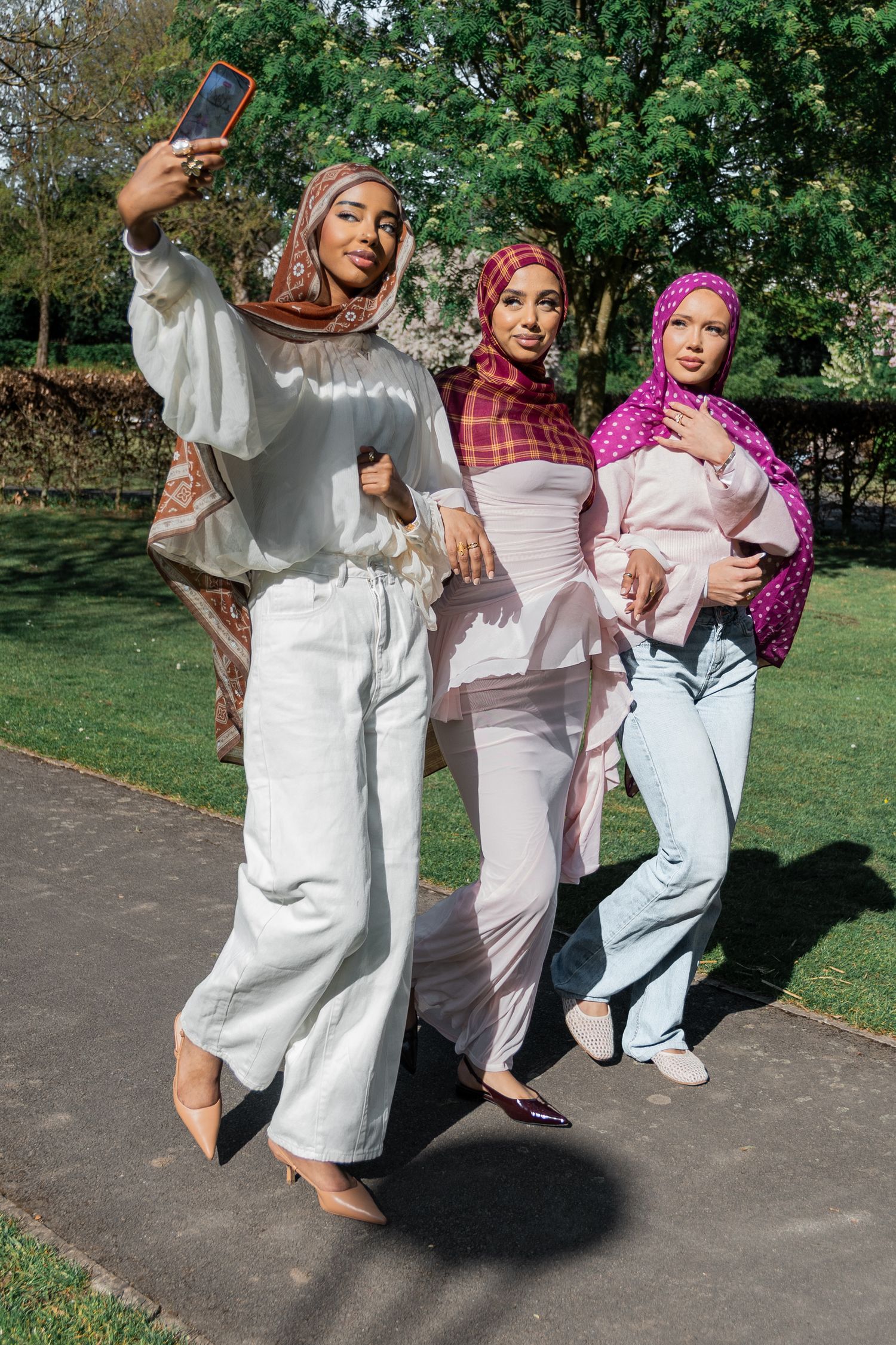 Three Muslim women in colorful hijabs take a selfie together in a sunny park setting.