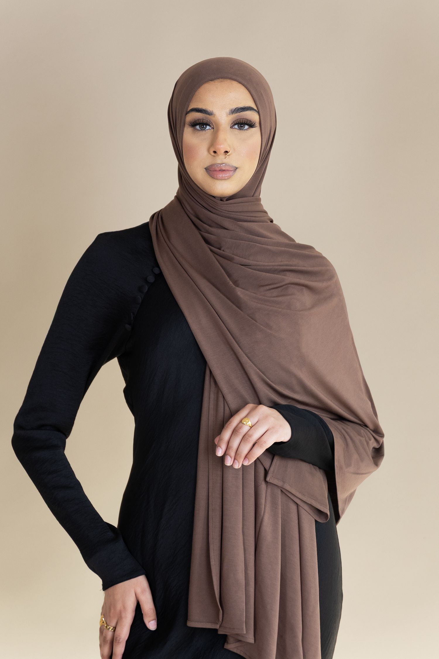 Person wearing a black long-sleeved top and brown hijab poses elegantly against neutral background.