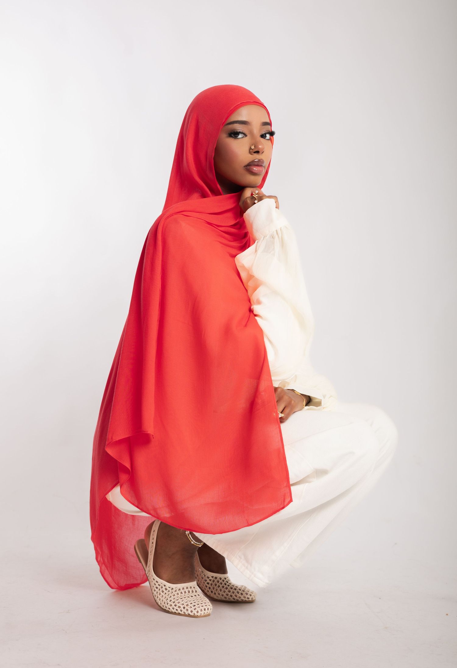 A woman wearing a vibrant red hijab and white outfit poses gracefully in a studio setting with neutral background.