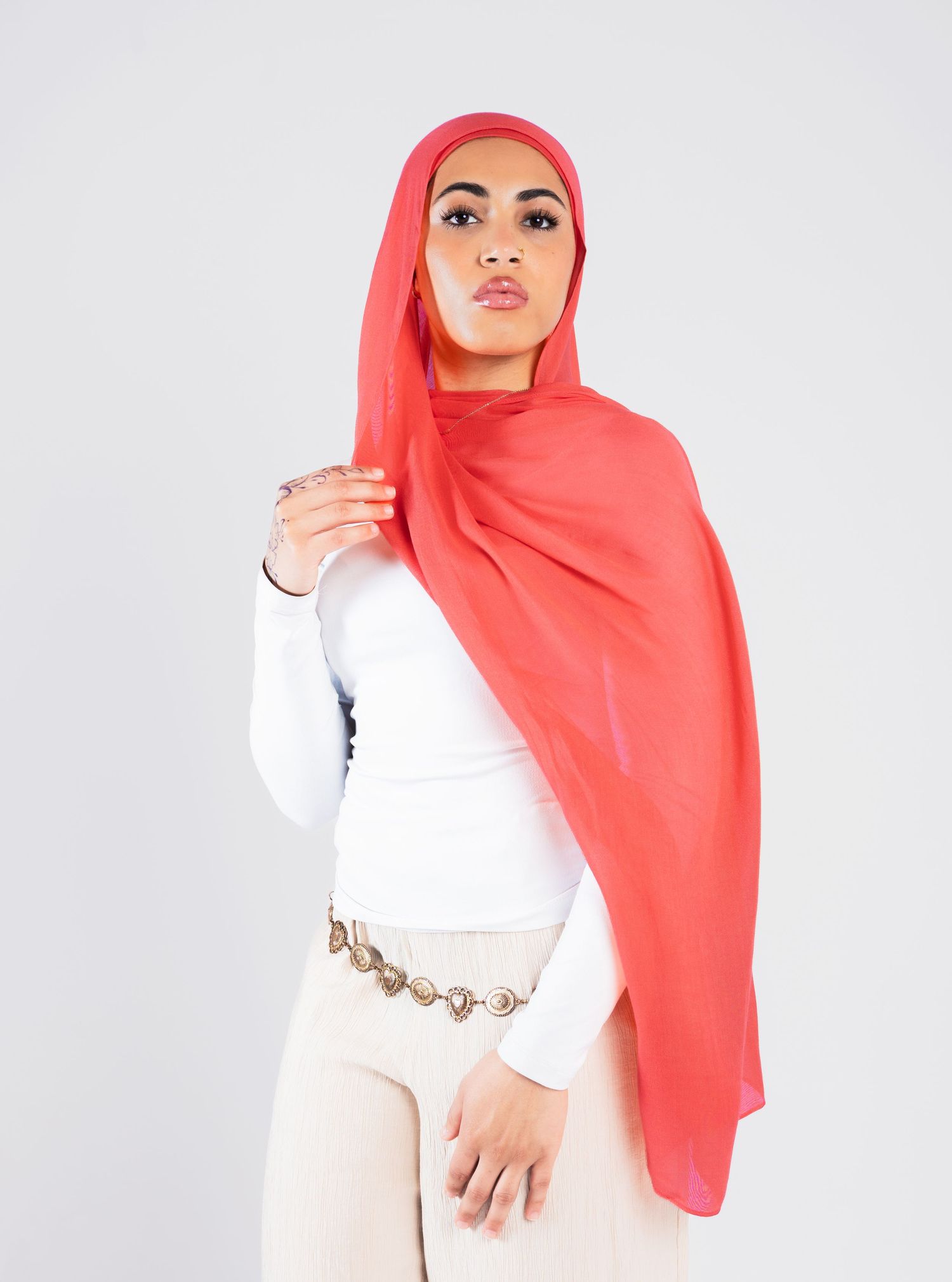 Woman wearing a coral red hijab and white top with a gold chain belt against a light gray background.