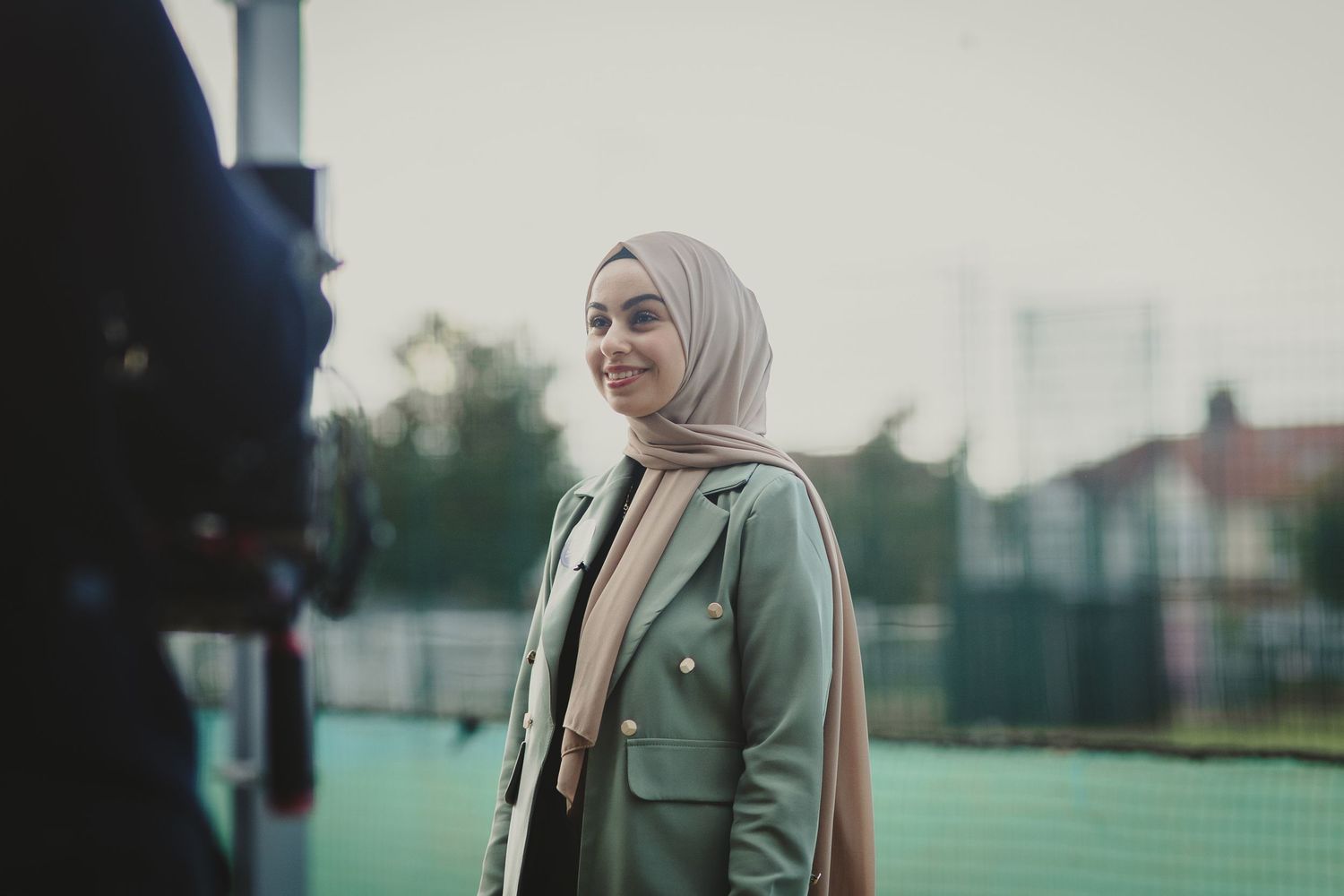 Athletic woman in hijab and green jacket stands outdoors in evening light on sports field.