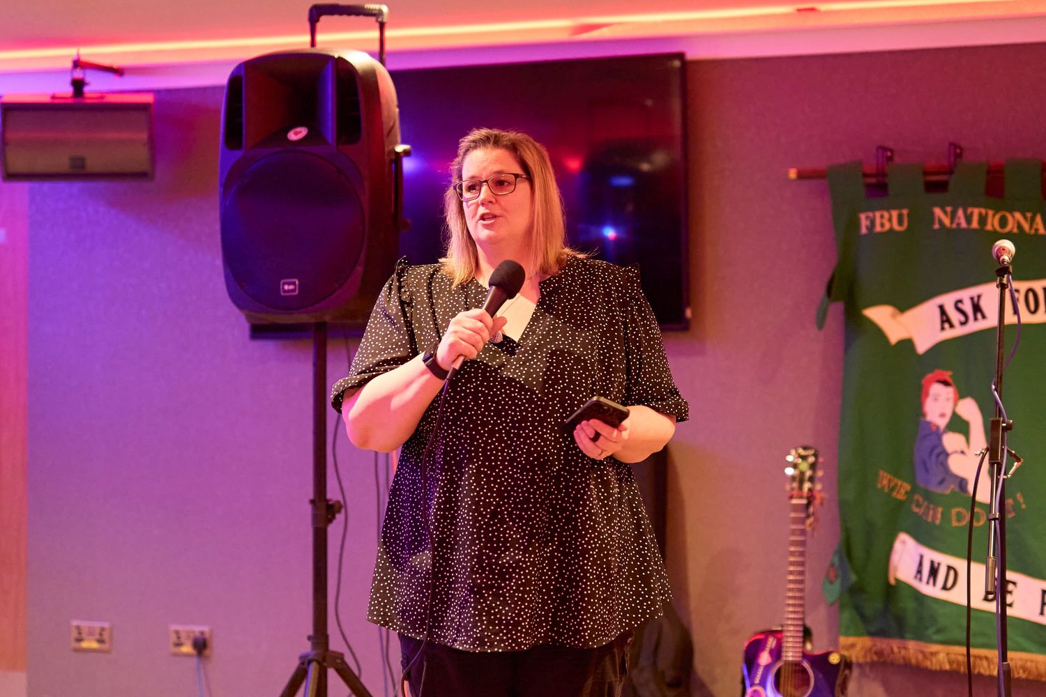 A performer in a polka dot dress stands on stage with a microphone in front of a large speaker and green banner.