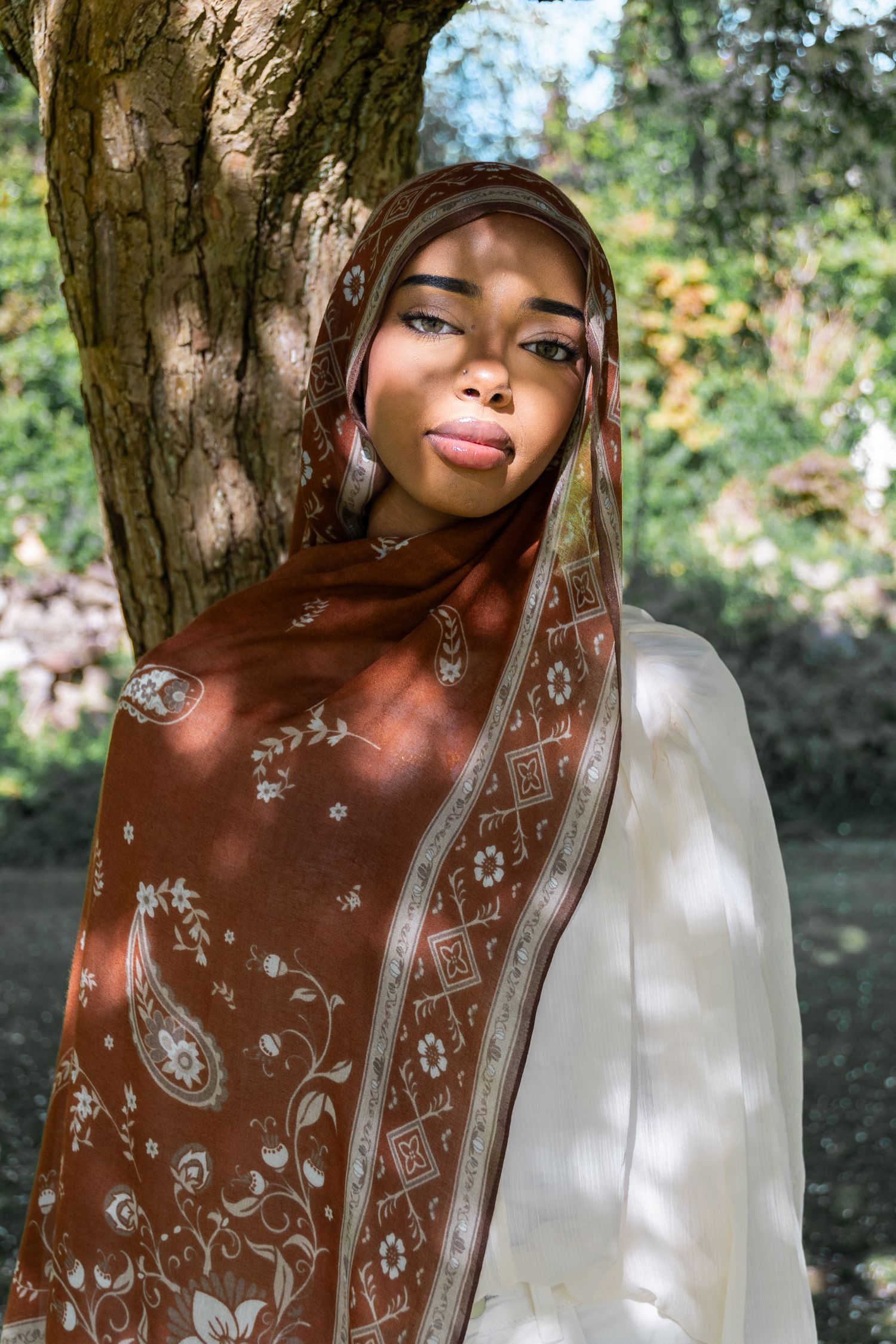 A young Black woman wearing a brown floral hijab and white dress poses gracefully beside a tree in a sunlit garden.