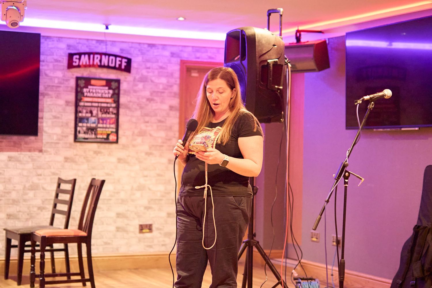 A performer stands on stage holding a microphone in a club setting with purple lighting and brick walls.