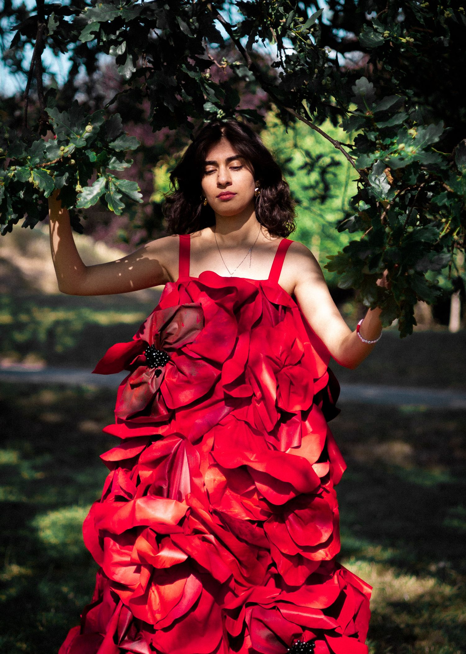 A dramatic red ruffled gown flows like rose petals against lush green foliage in an artistic outdoor portrait.