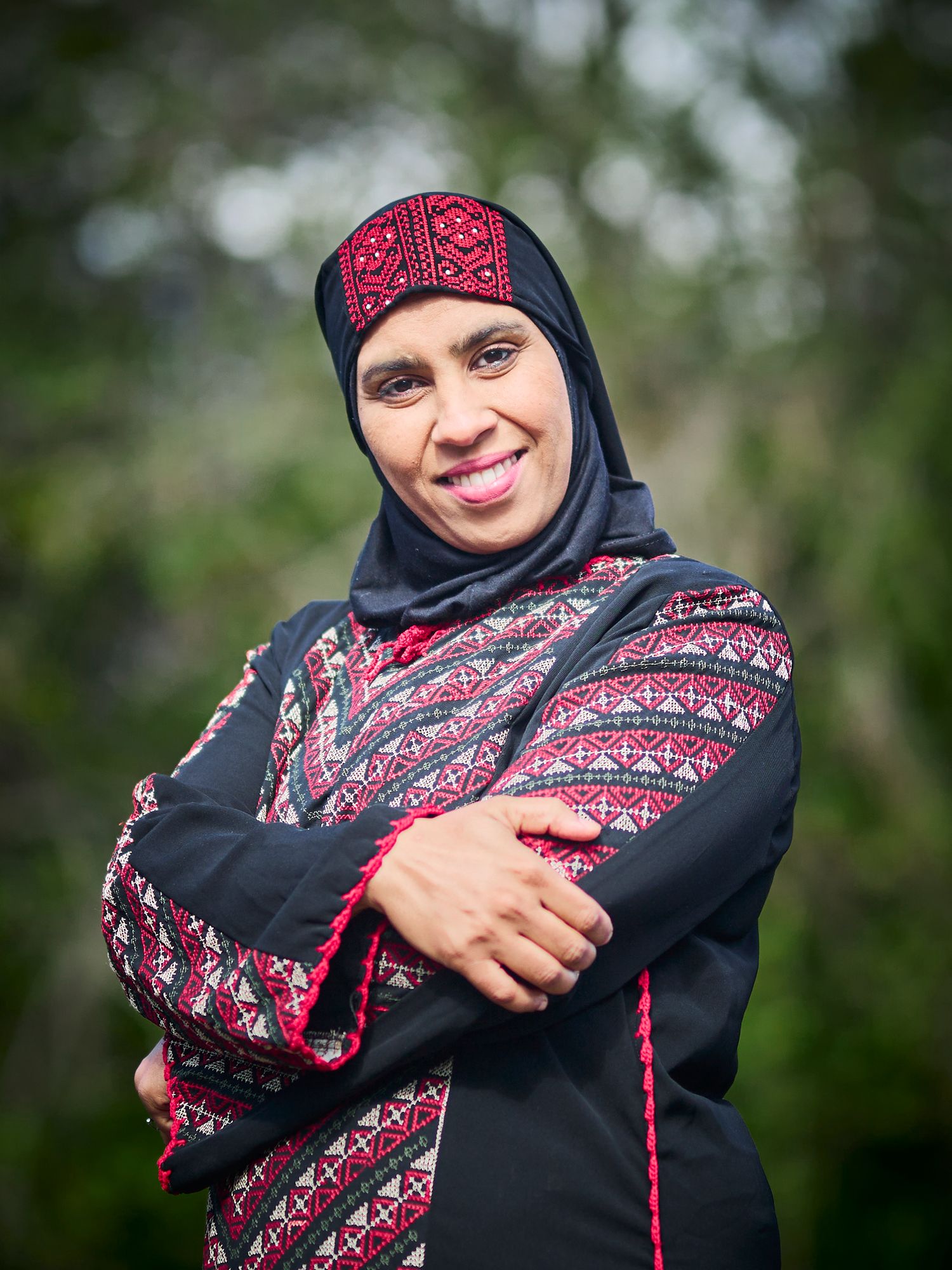 Woman in traditional black and red patterned dress and hijab poses with crossed arms.