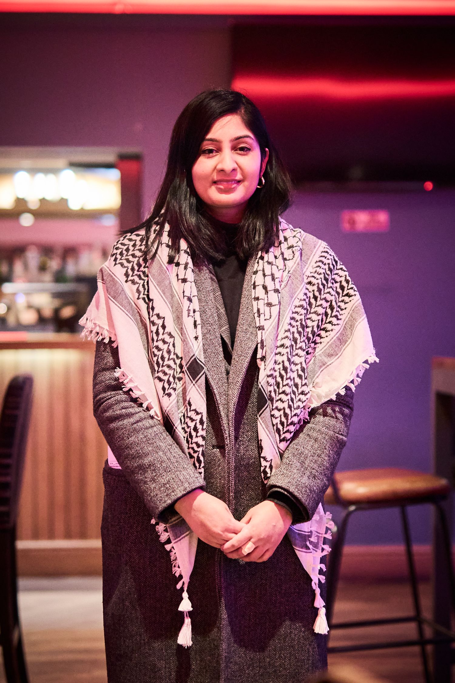 Woman wearing patterned keffiyeh scarf over gray sweater smiles in indoor setting.