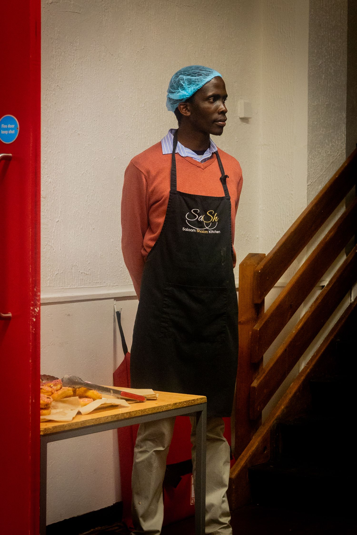 Restaurant worker in black apron and blue cap standing near wooden stairs.
