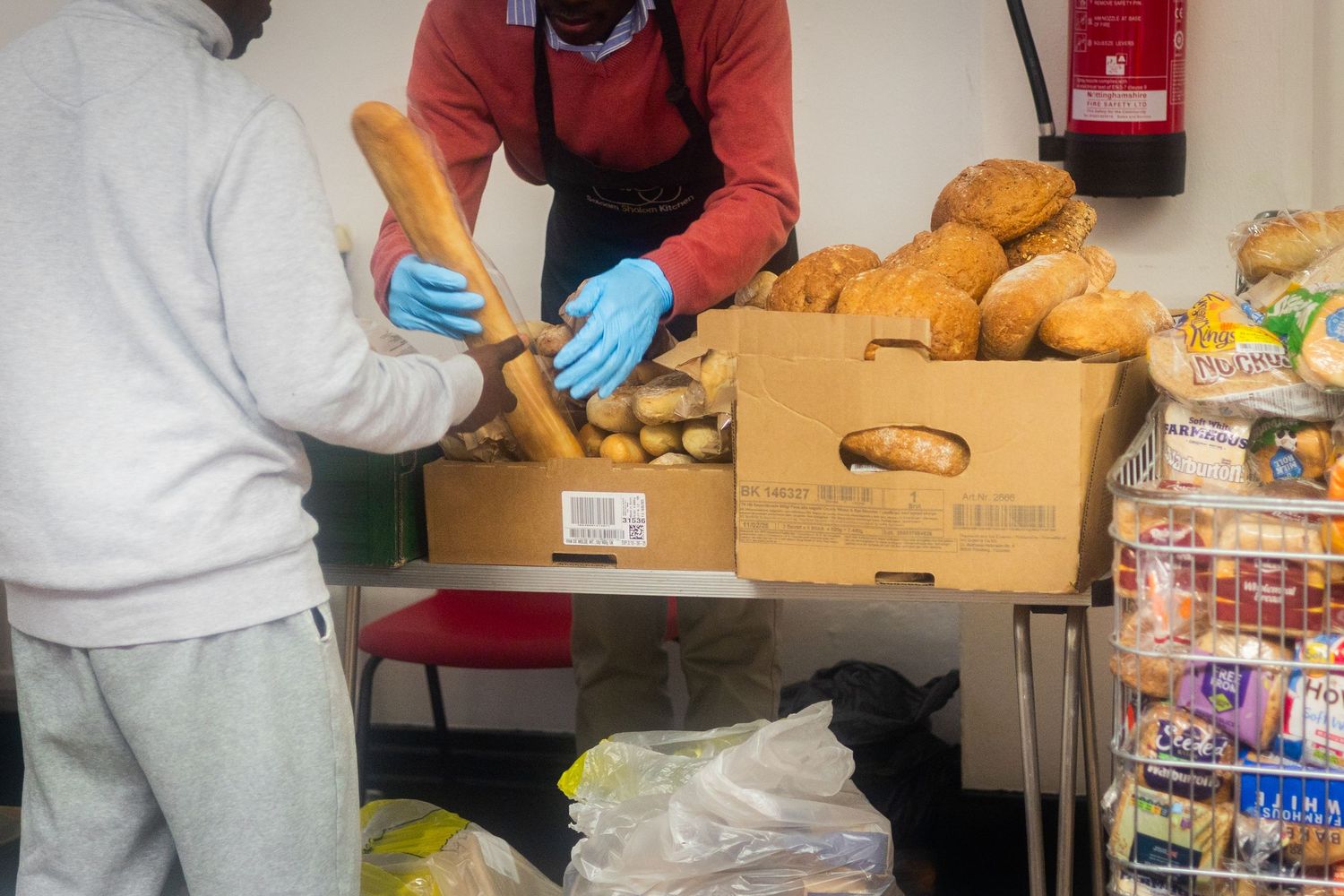 Volunteers in protective gloves sort and pack food items into cardboard boxes at a distribution center.