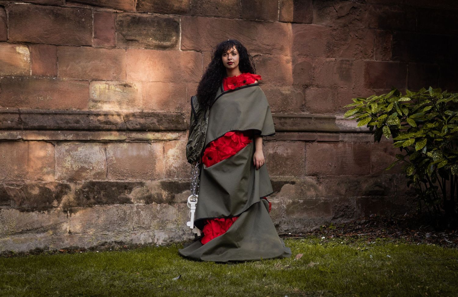 A figure wrapped in a gray and red blanket stands against a rustic stone wall with green foliage.