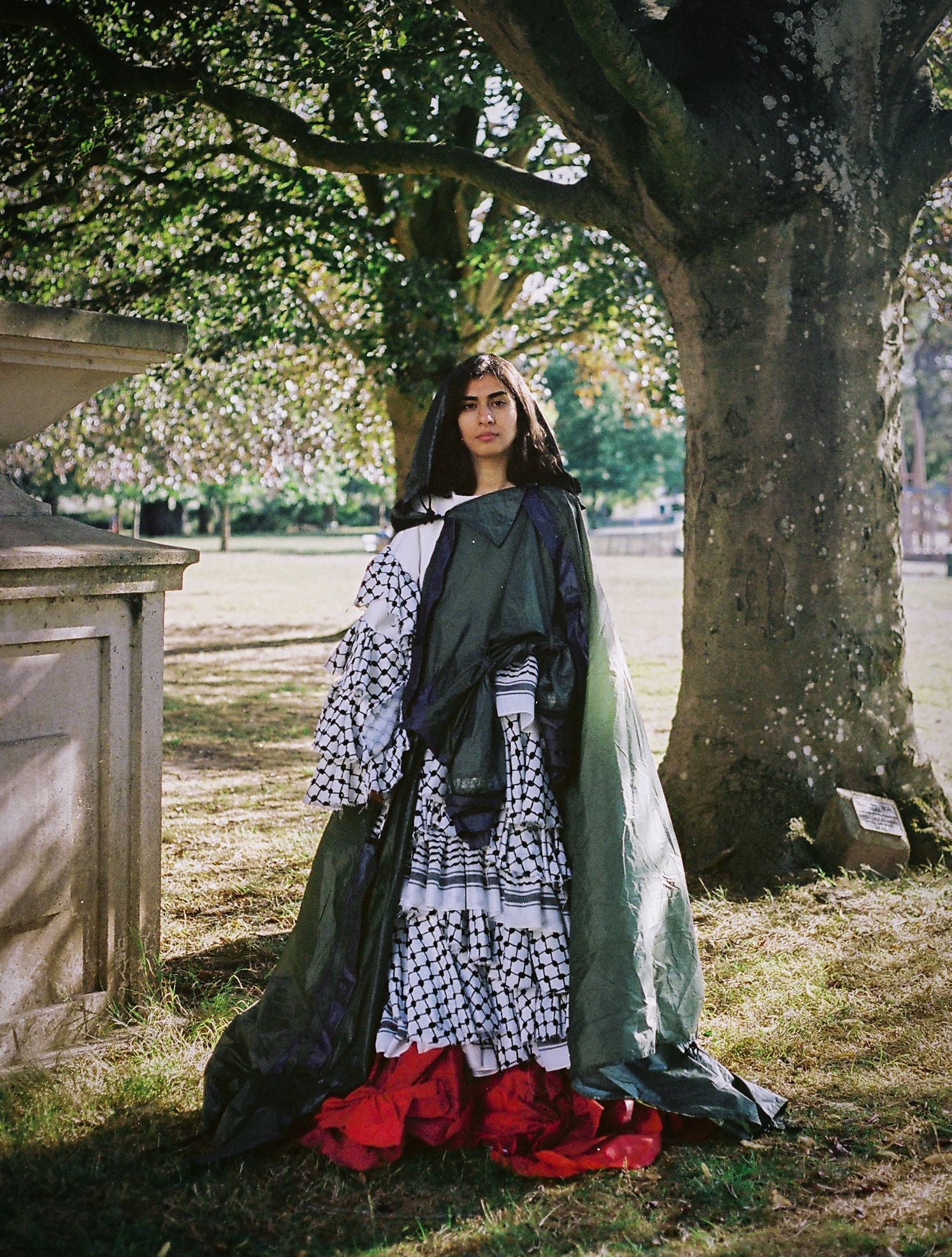 Fashionable outfit featuring black coat, checkered dress and red boots under a large tree in a garden setting.