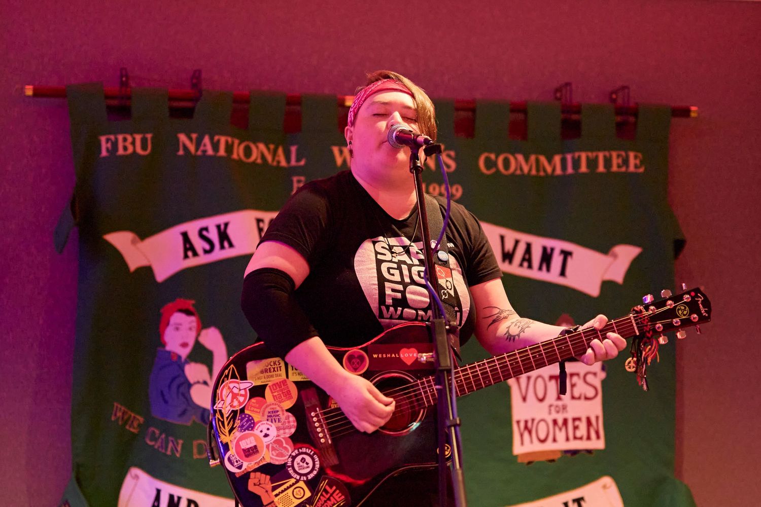 A performer plays guitar and sings on stage with a pink-tinted backdrop featuring political banners and signage.