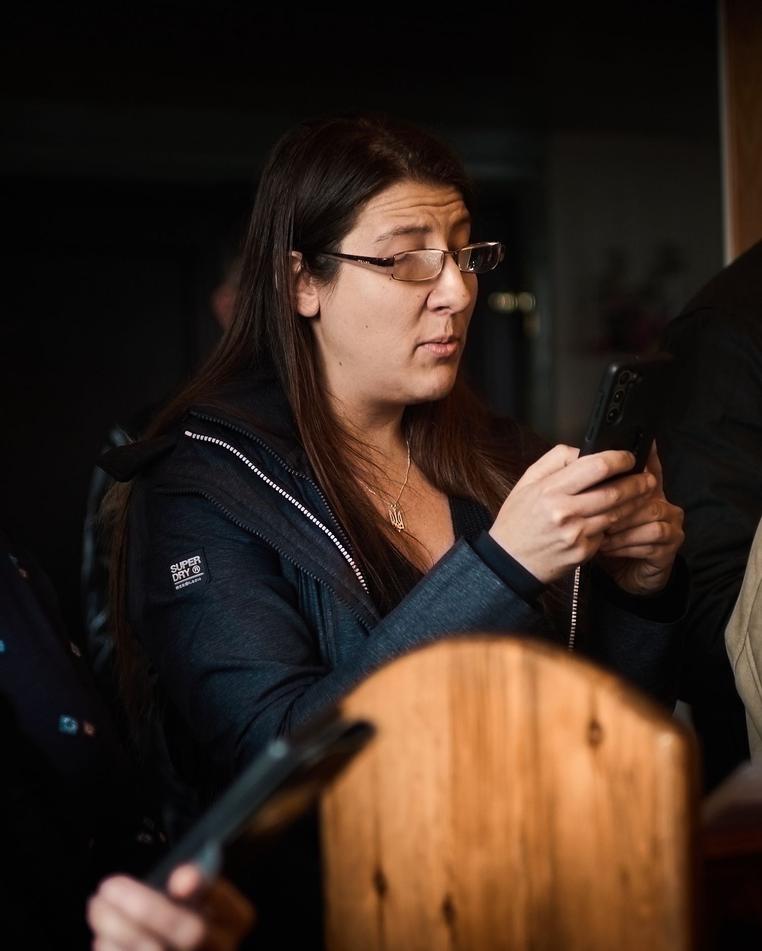Woman with glasses and dark hair looking at her phone while holding items indoors.