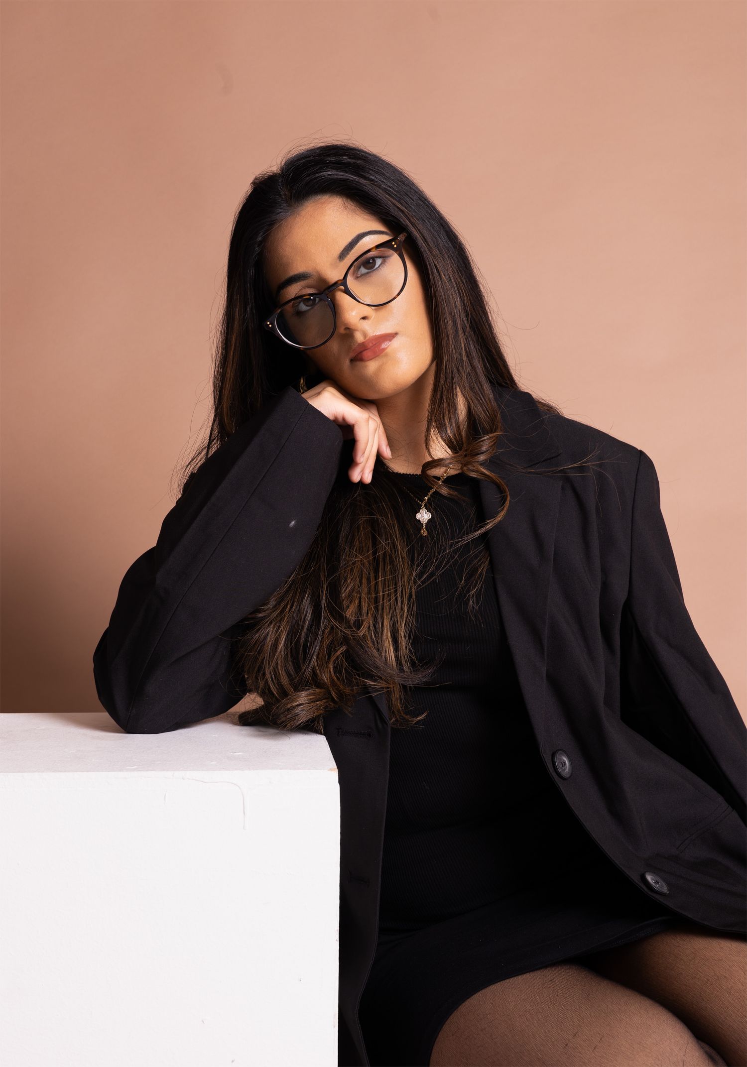 Young woman in black blazer and round glasses poses elegantly against a warm beige studio background.