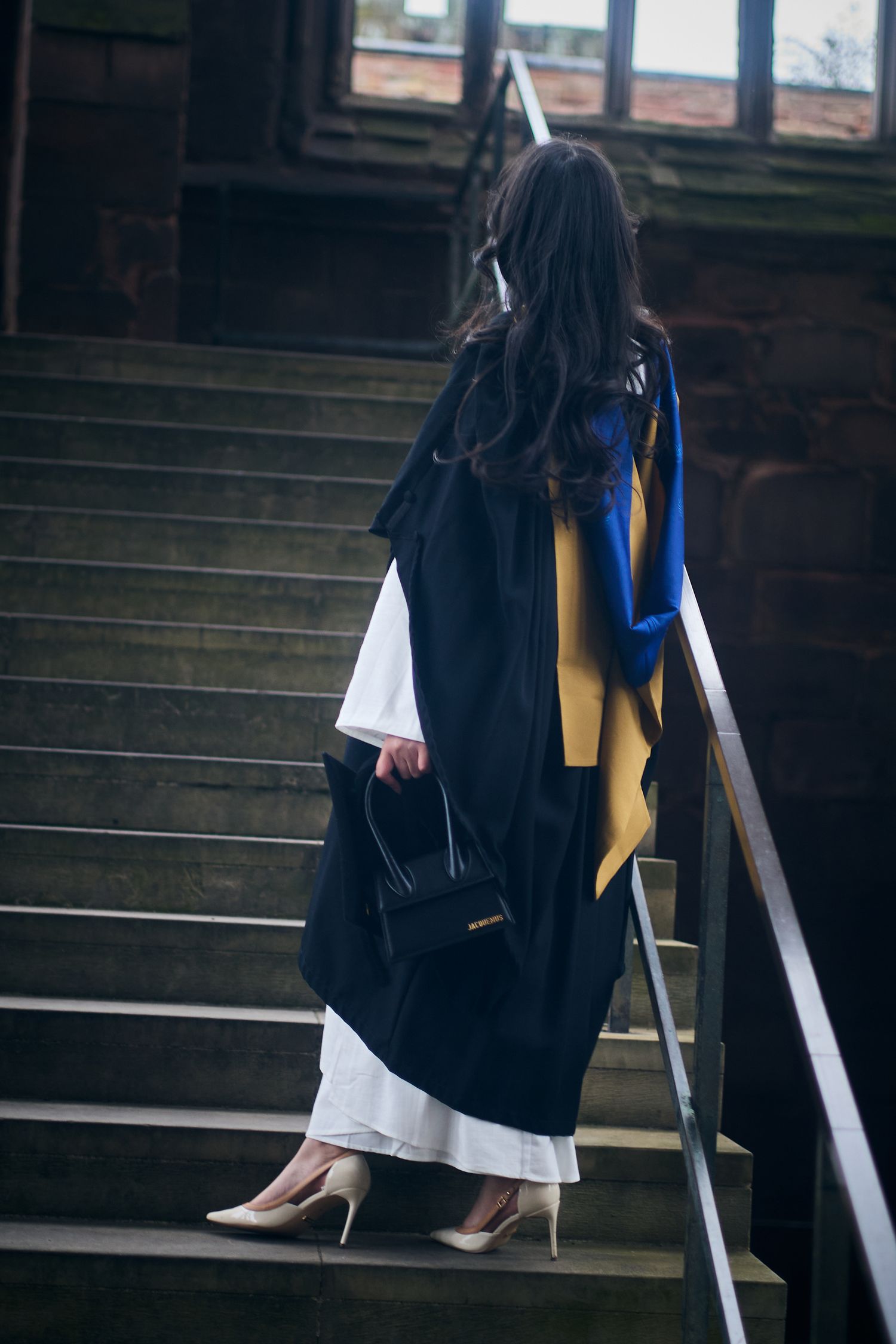 A person in a black graduation gown walks down wooden stairs while holding academic regalia.