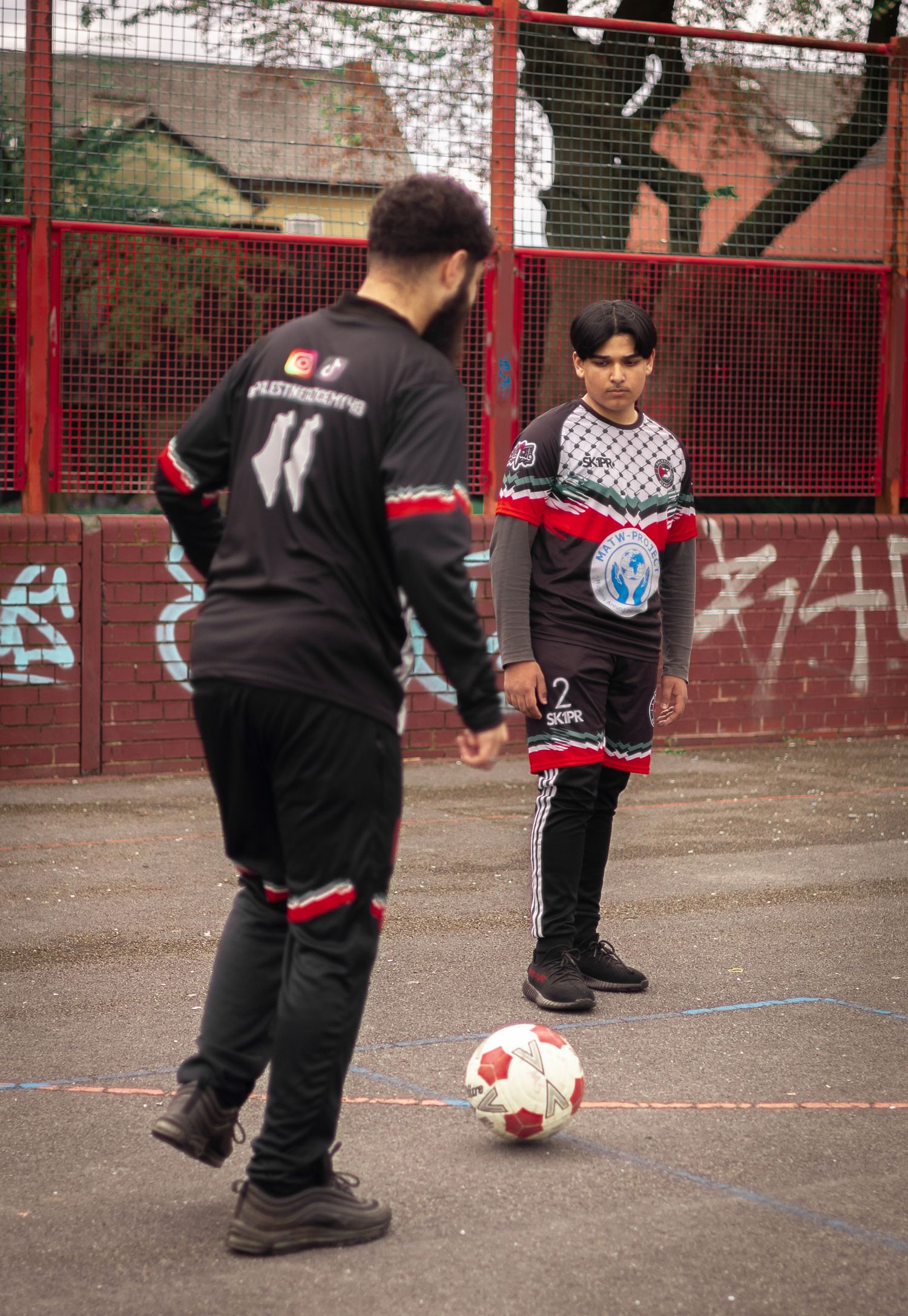 Two players in black uniforms practice soccer skills on an outdoor court with red fencing.
