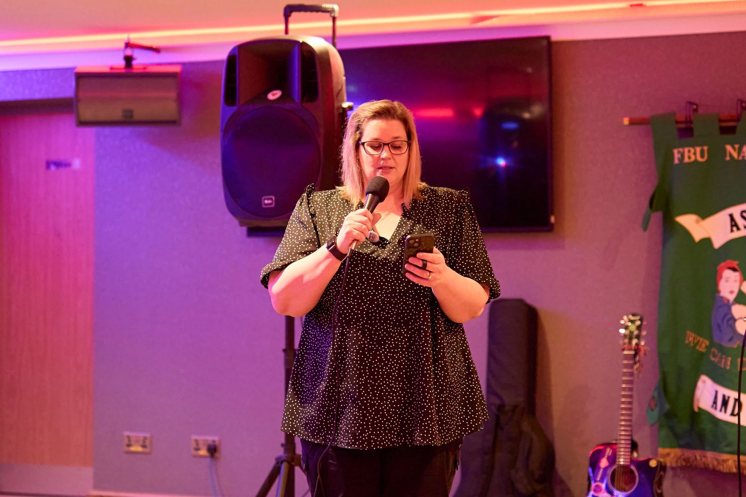 A performer in a polka dot dress holds a microphone on stage with pink and purple lighting and audio equipment visible.