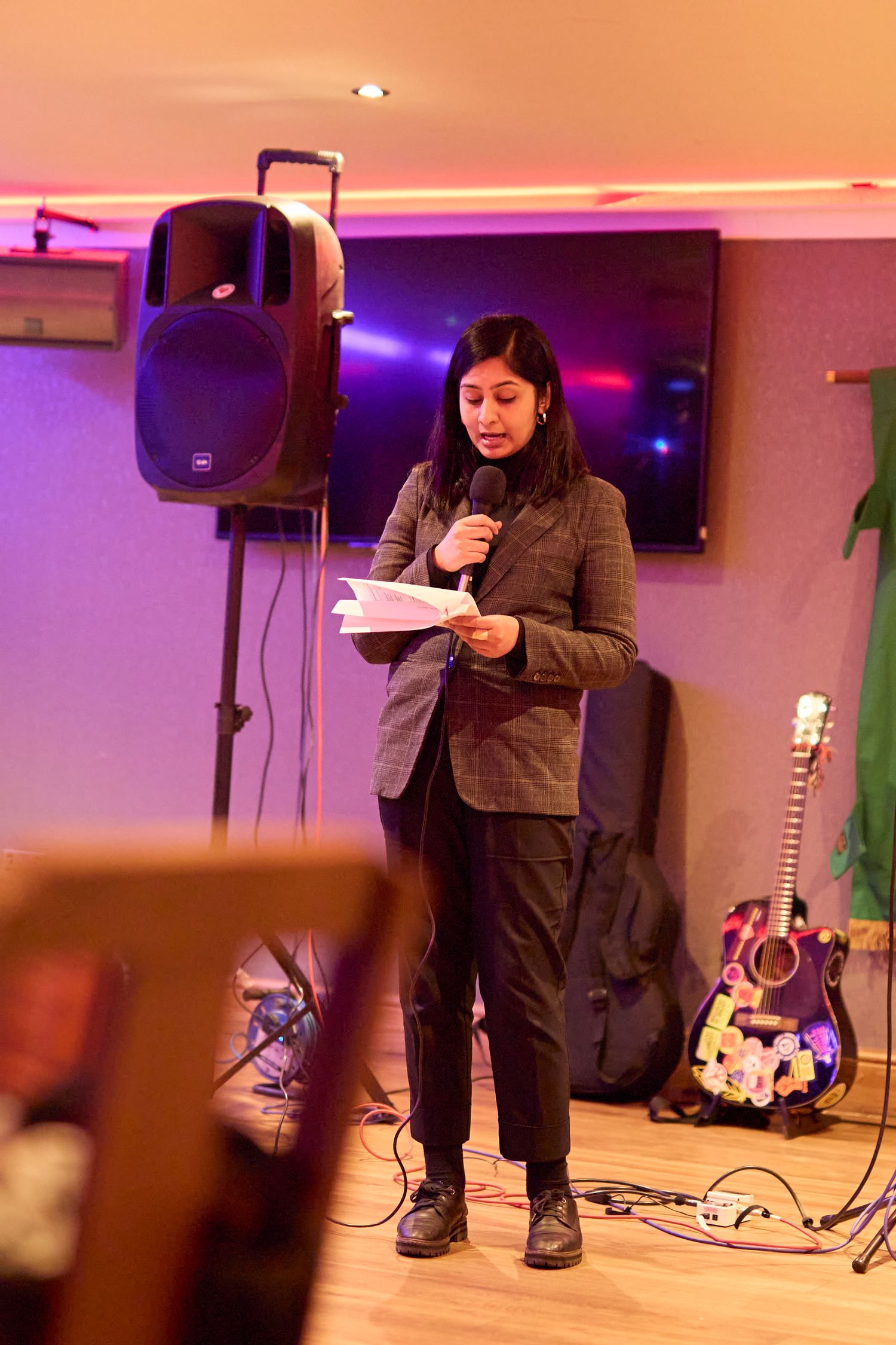 A speaker stands at a microphone in a room with purple lighting and musical equipment including a guitar in the background.
