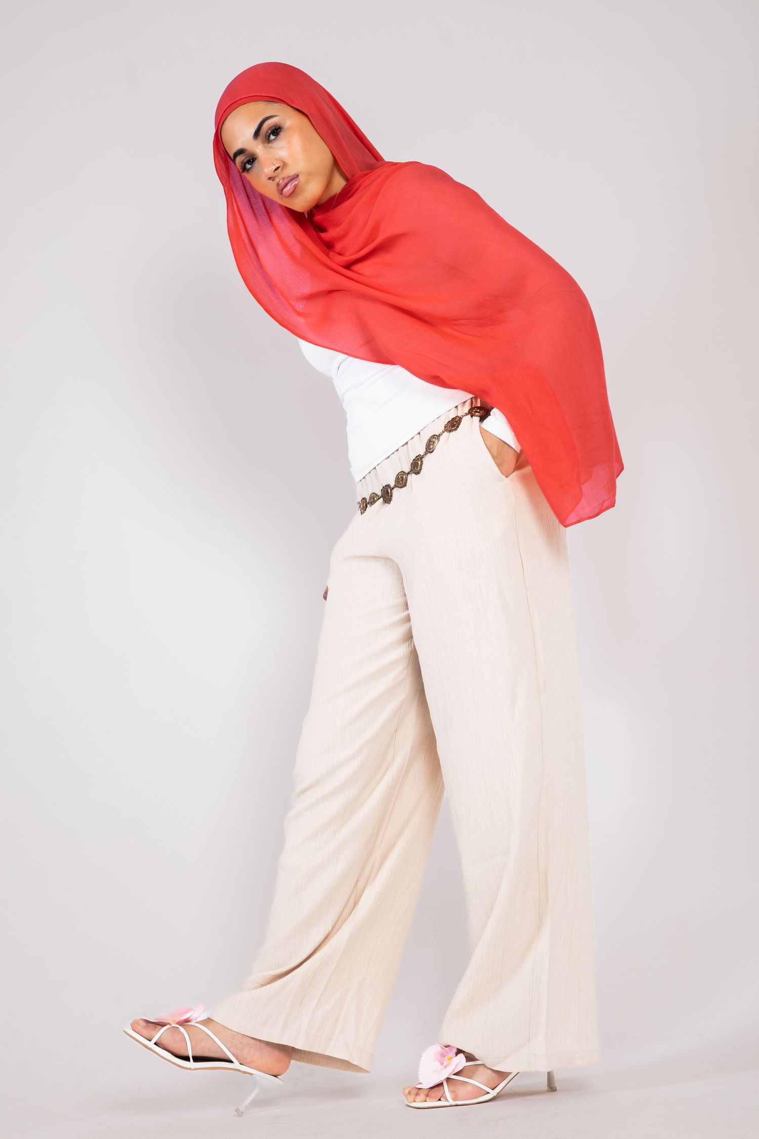Woman wearing red hijab, white top, cream wide-leg pants, and white sandals posing in a studio.