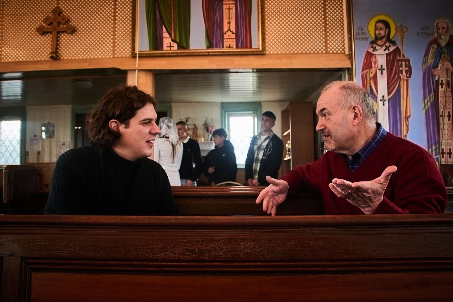 Two people engaged in conversation inside a traditional church setting with religious icons visible.