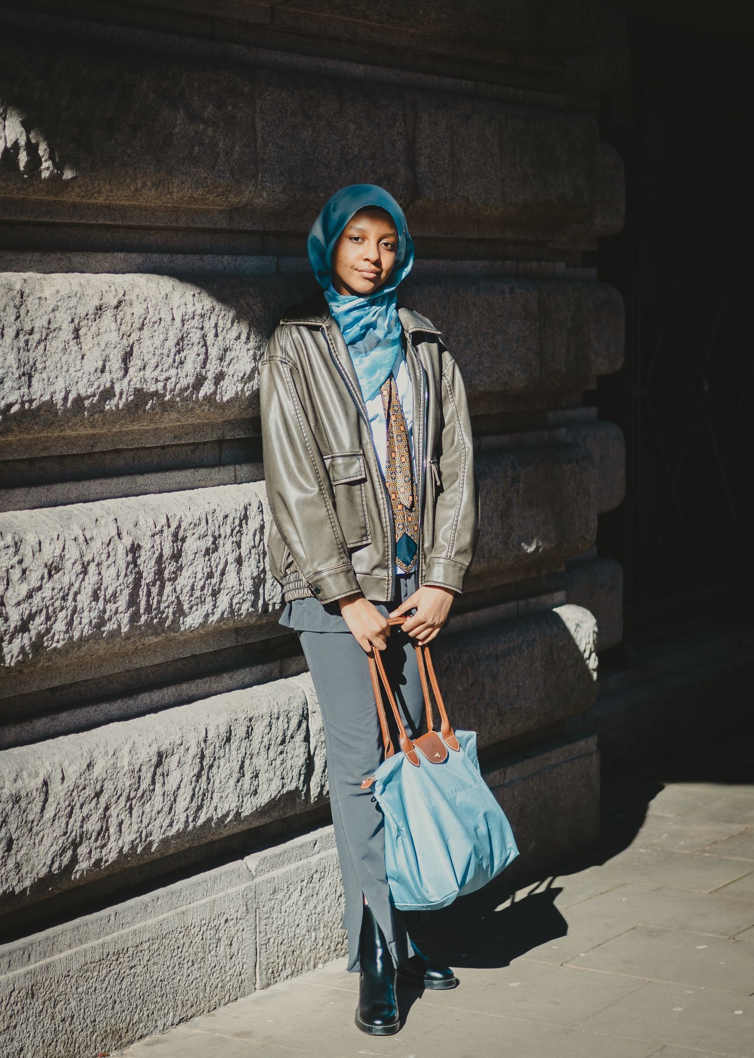Urban fashion shoot with light brown jacket and blue accessories against stone wall texture.