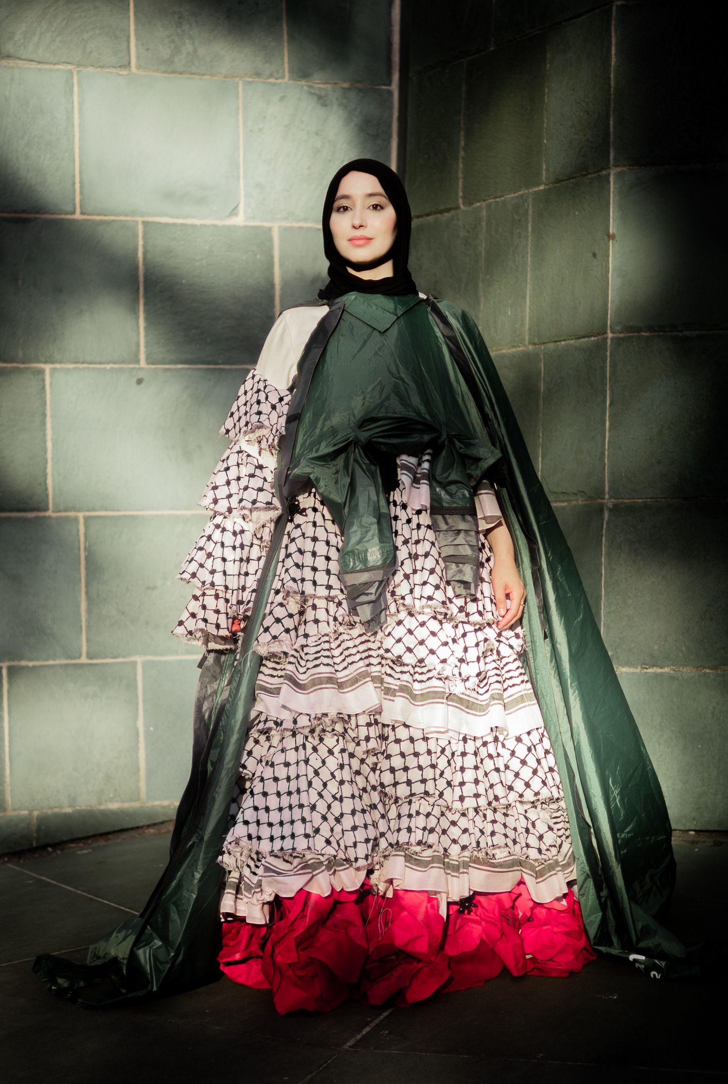 A person wearing a dramatic green cape over a patterned white dress with red ruffles stands against stone walls in moody lighting.