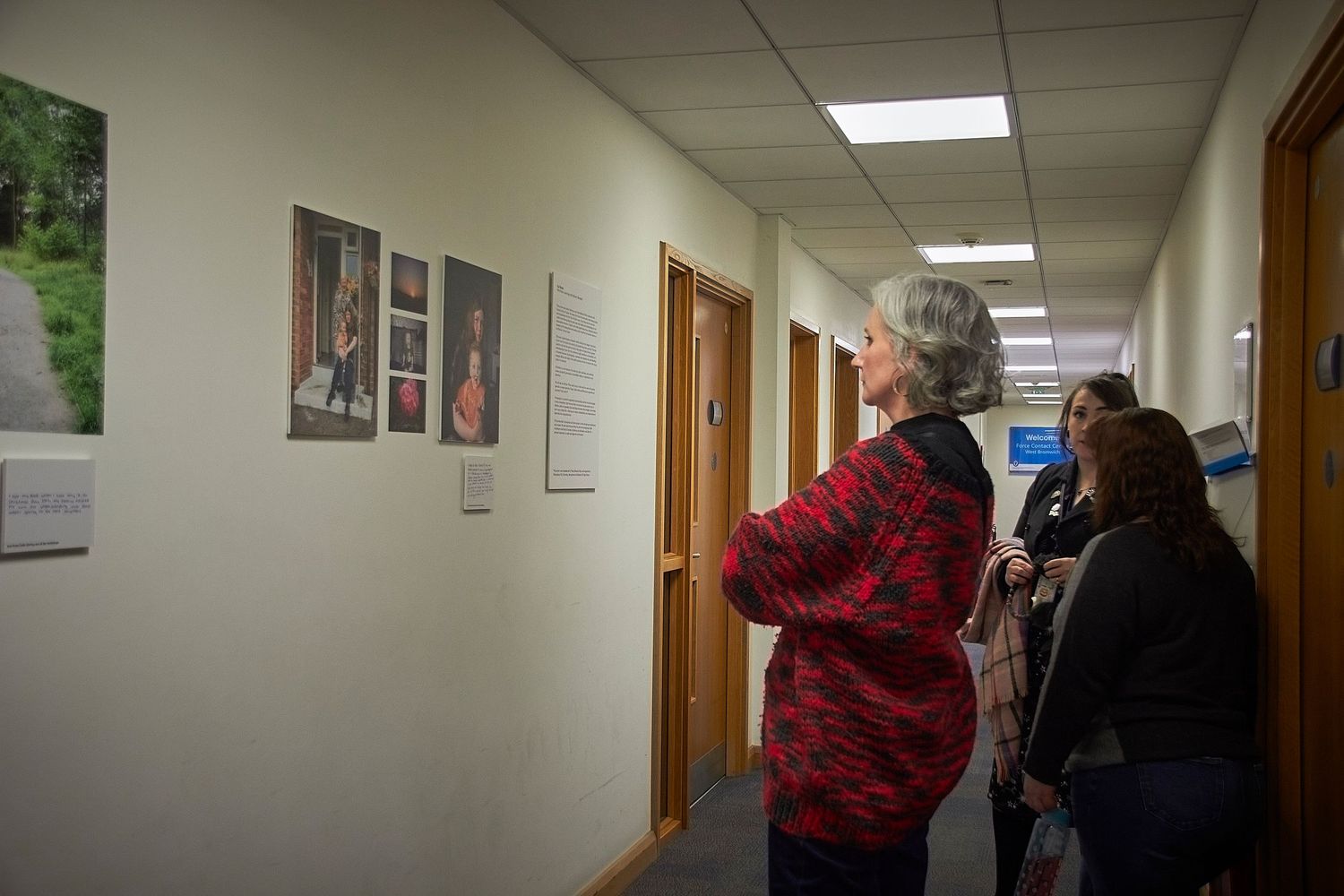 People viewing photographs displayed on a hallway wall in an office or institutional building.