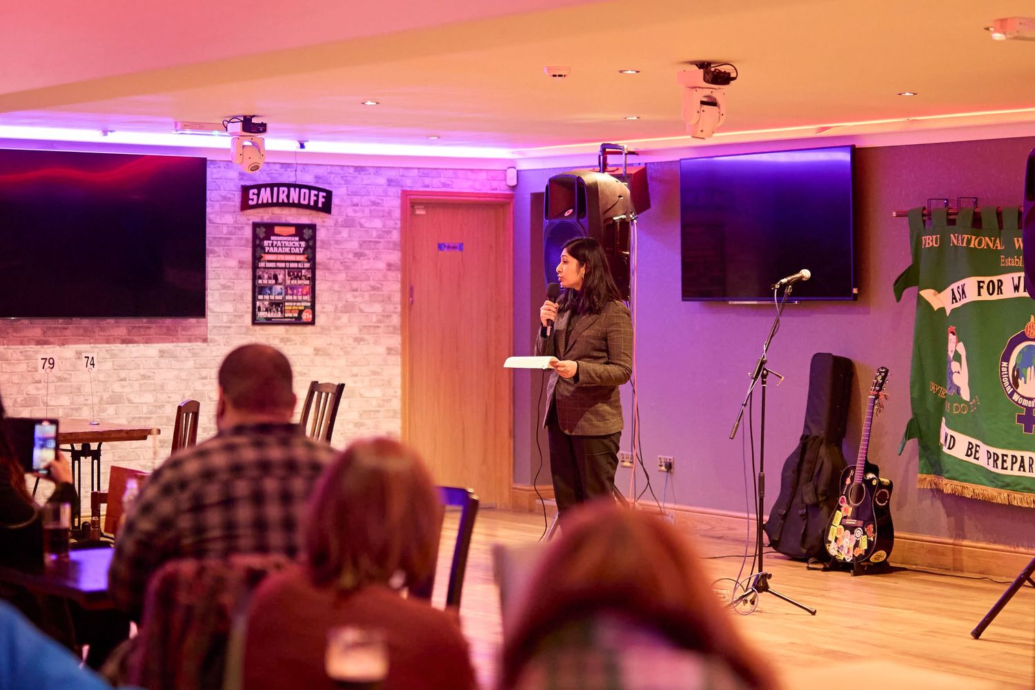 A bar venue with purple lighting features TV screens, brick walls, a microphone stand, and a green flag banner.