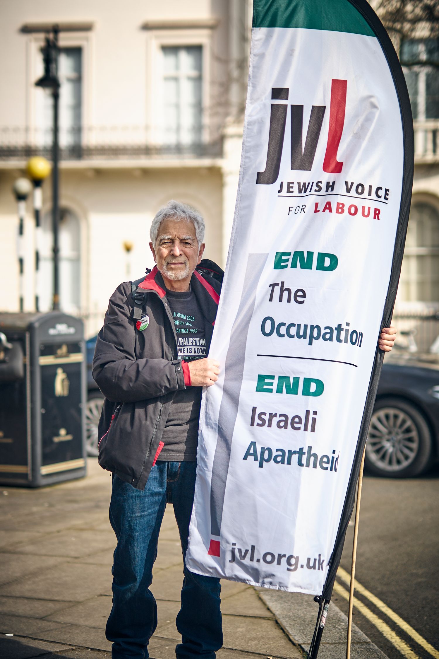 Protester stands next to Jewish Voice for Labour banner at street demonstration.