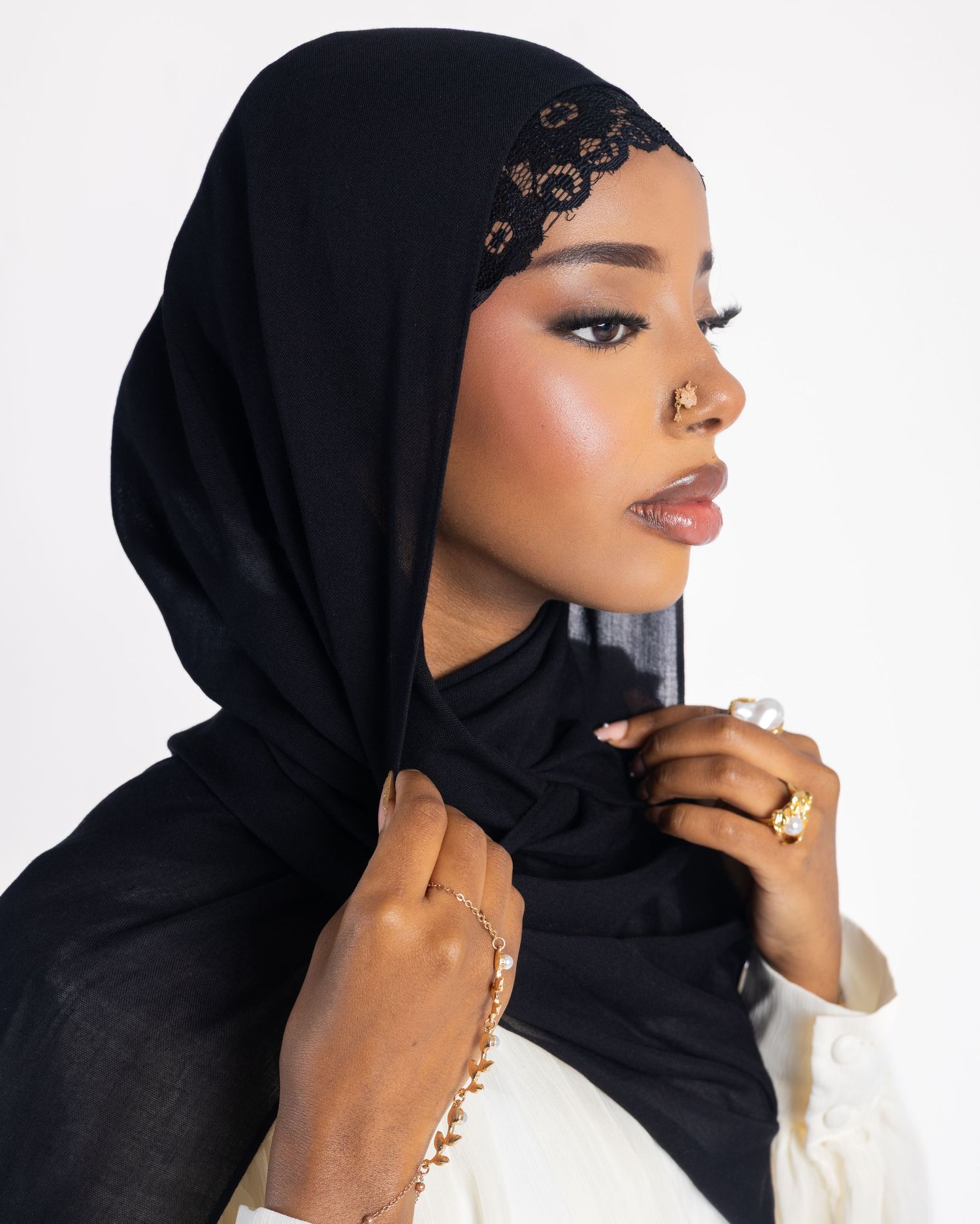 Woman wearing a black hijab with lace detail and gold jewelry, posing against a white background.
