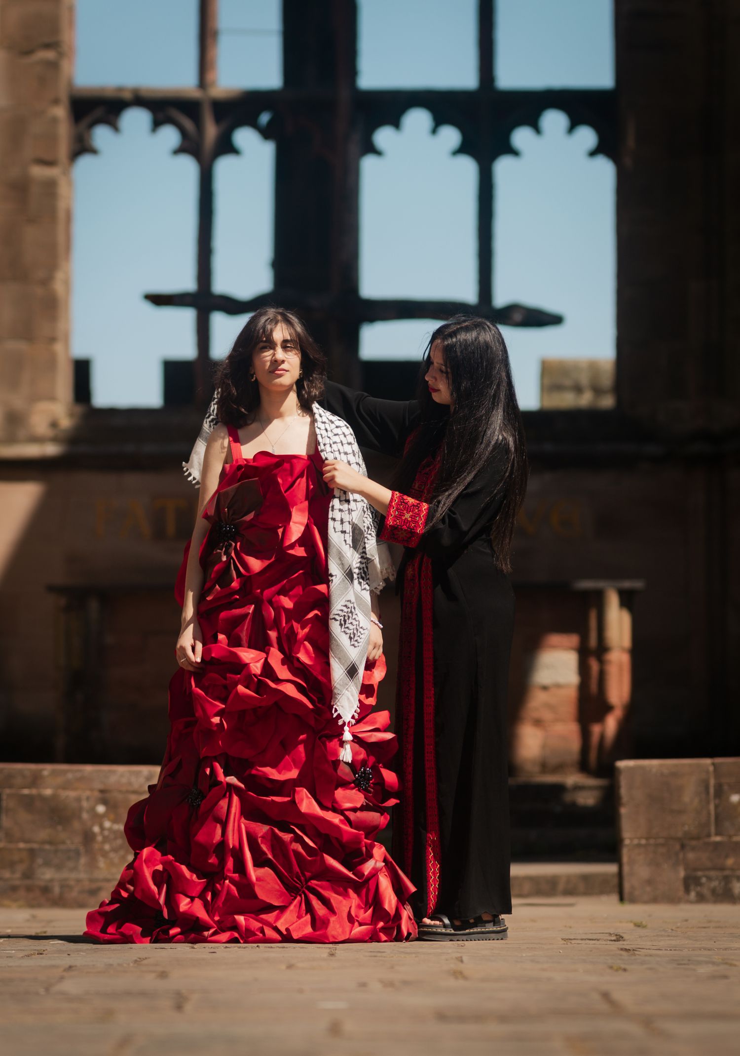 Fashion scene with red ruffled gown and styling assistant against gothic architecture.