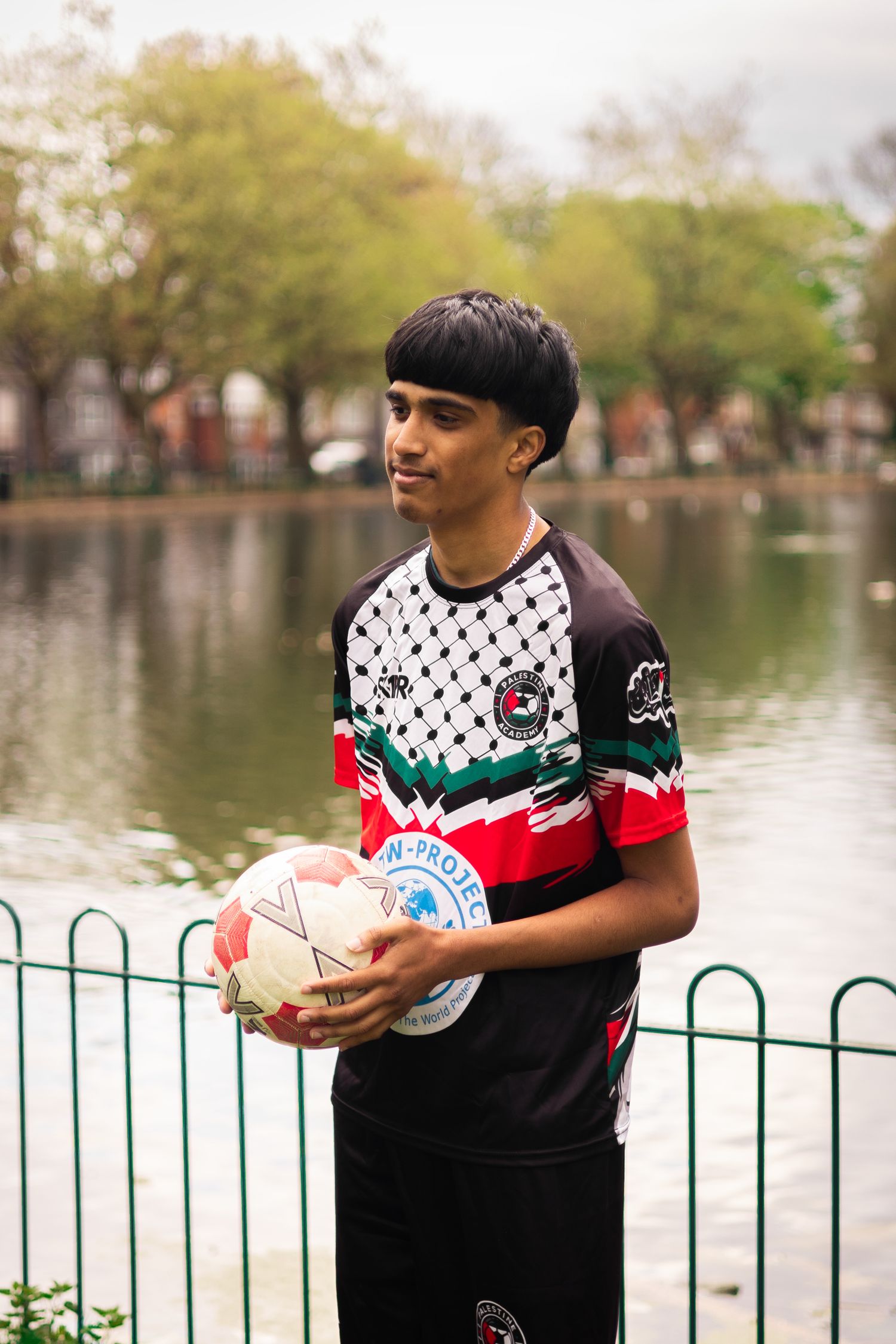 Player in stylish team uniform holds soccer ball while standing by pond with railing.