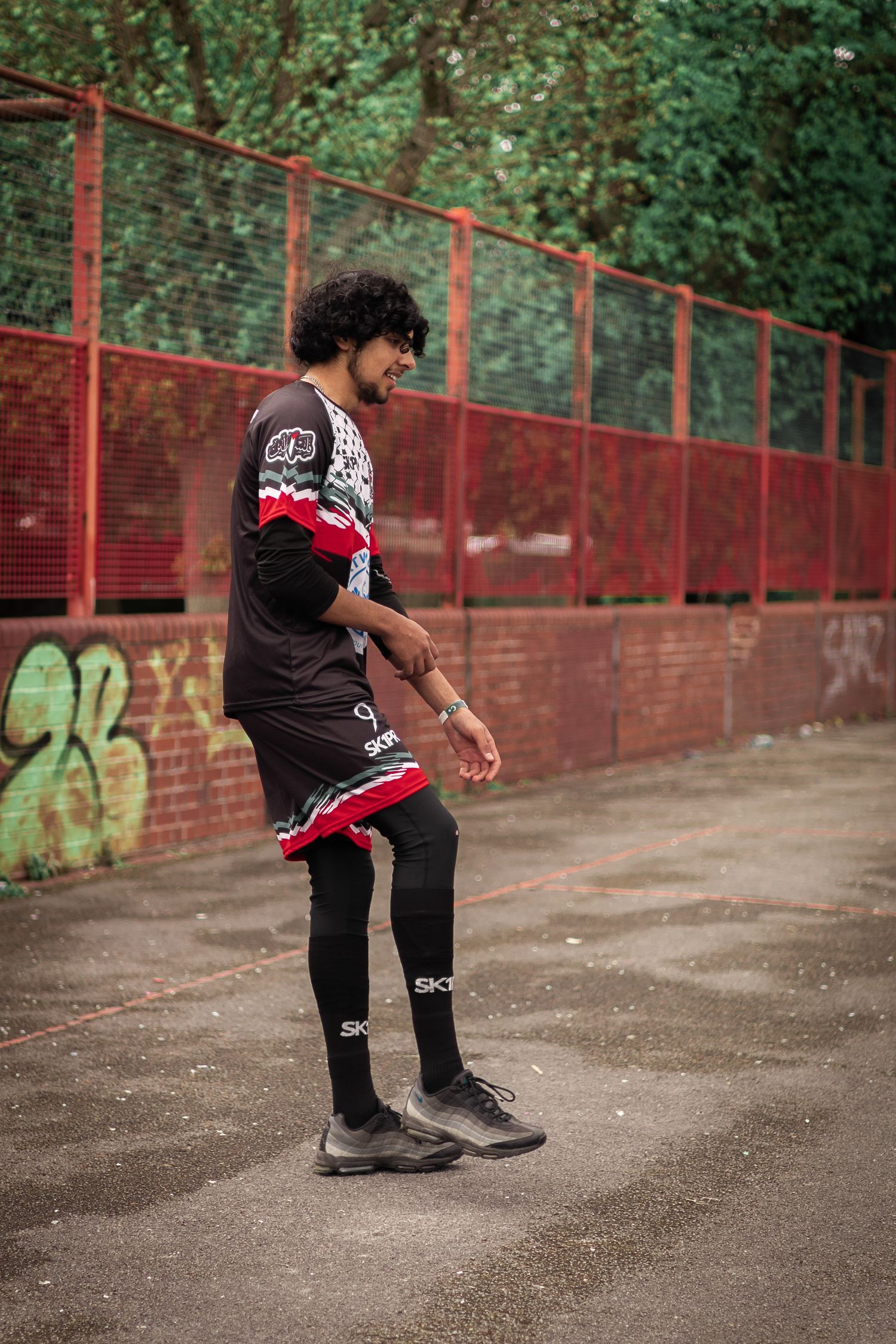 Player in black and red uniform stretching on outdoor court with red fence background.