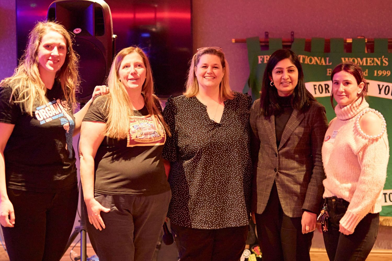 A group of five people smile together on stage with red lighting and signage visible in the background.