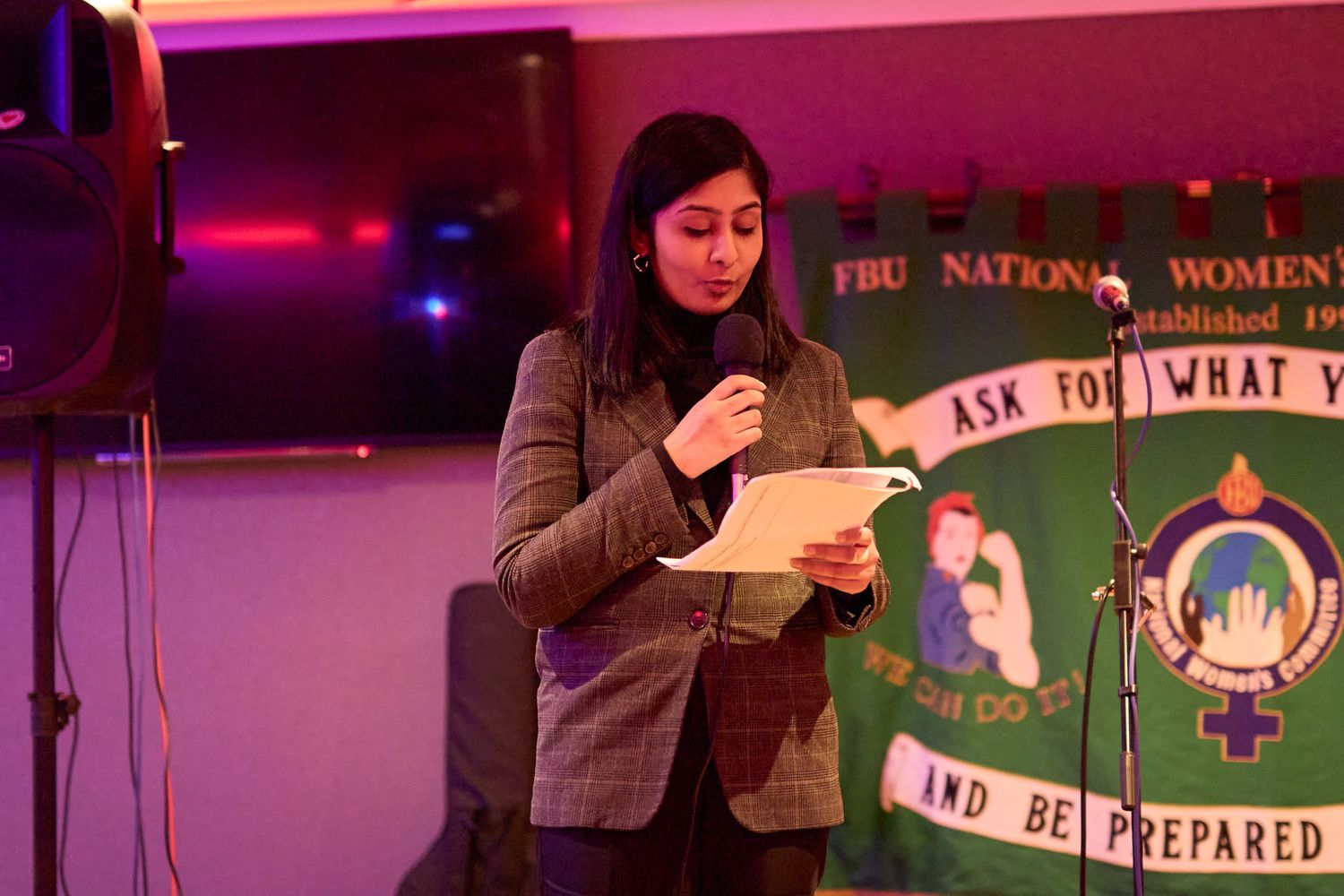 A speaker at a podium reads from notes under pink lighting with a banner visible in the background.