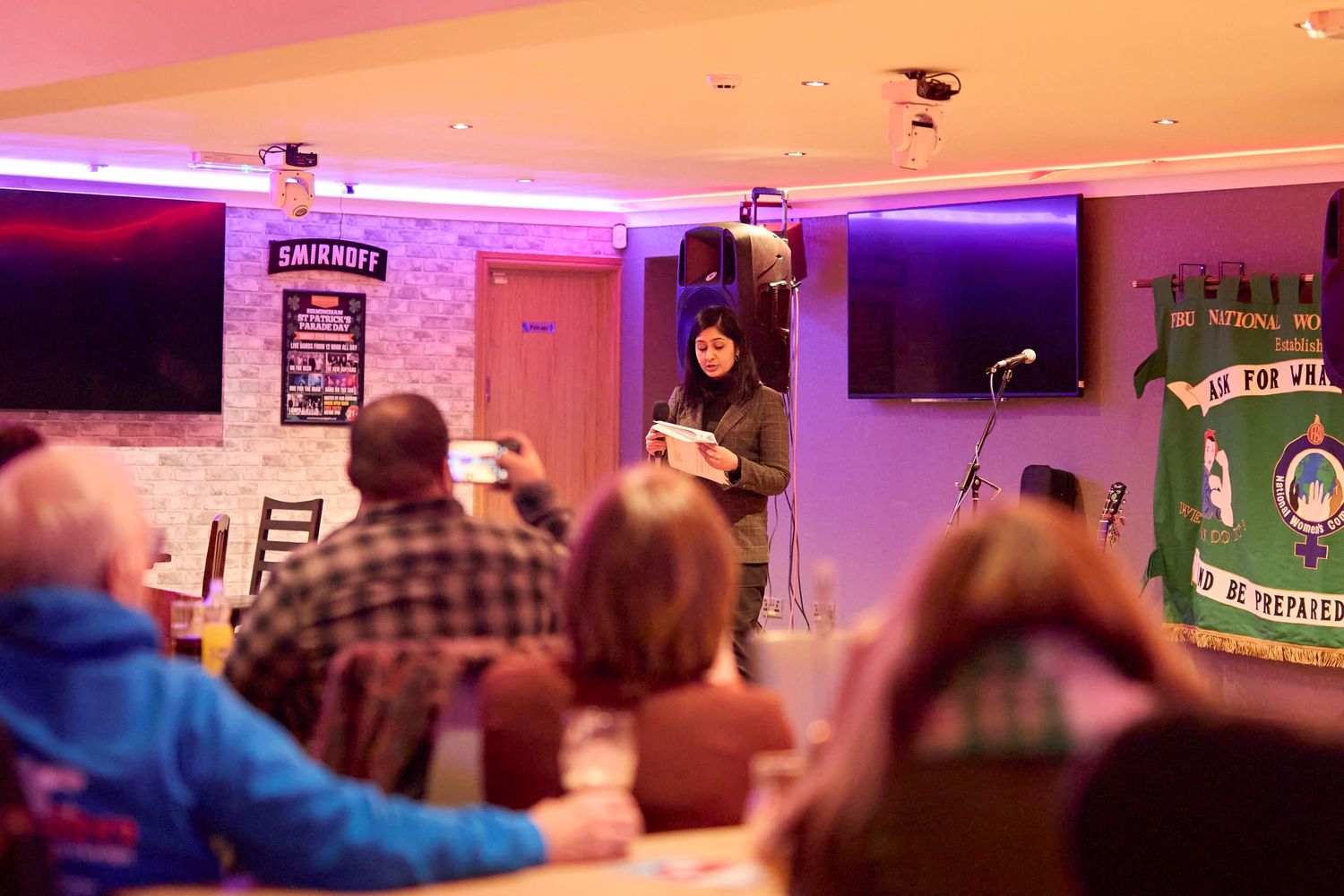 A panoramic view of a dimly lit bar or event space with purple and orange ambient lighting and guests mingling.