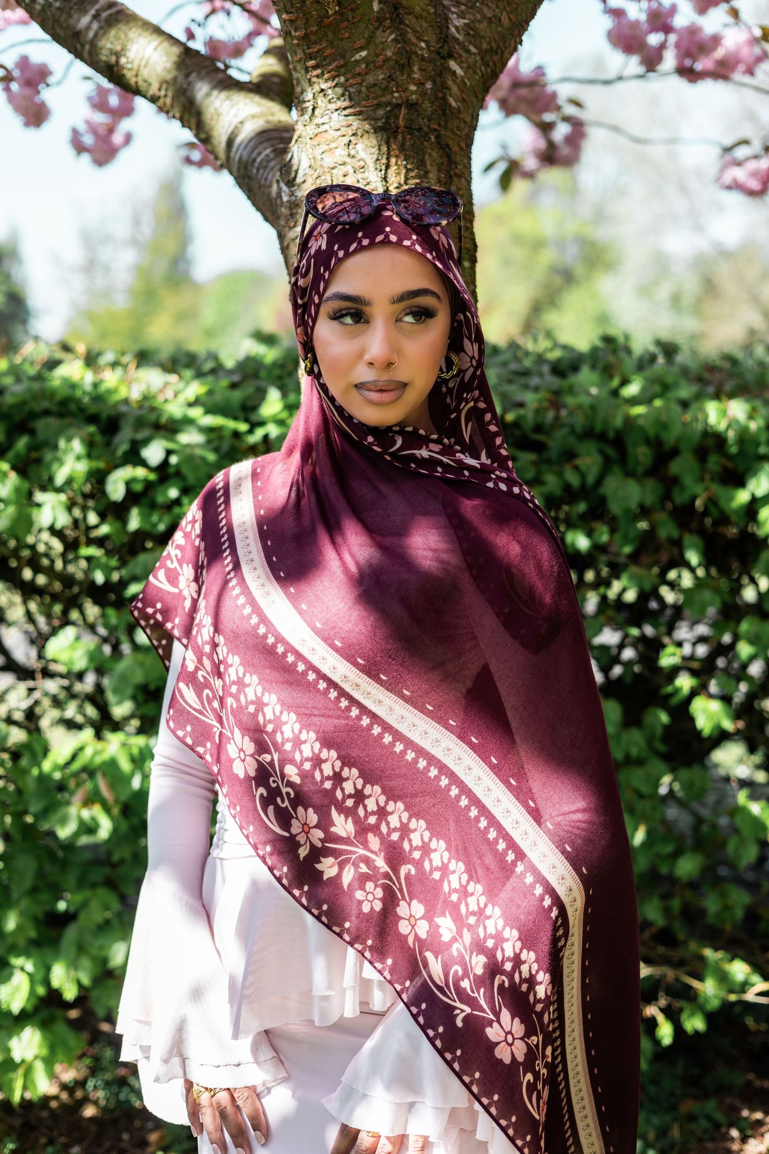 Woman in burgundy floral hijab stands before blooming tree and lush greenery in a serene outdoor setting.