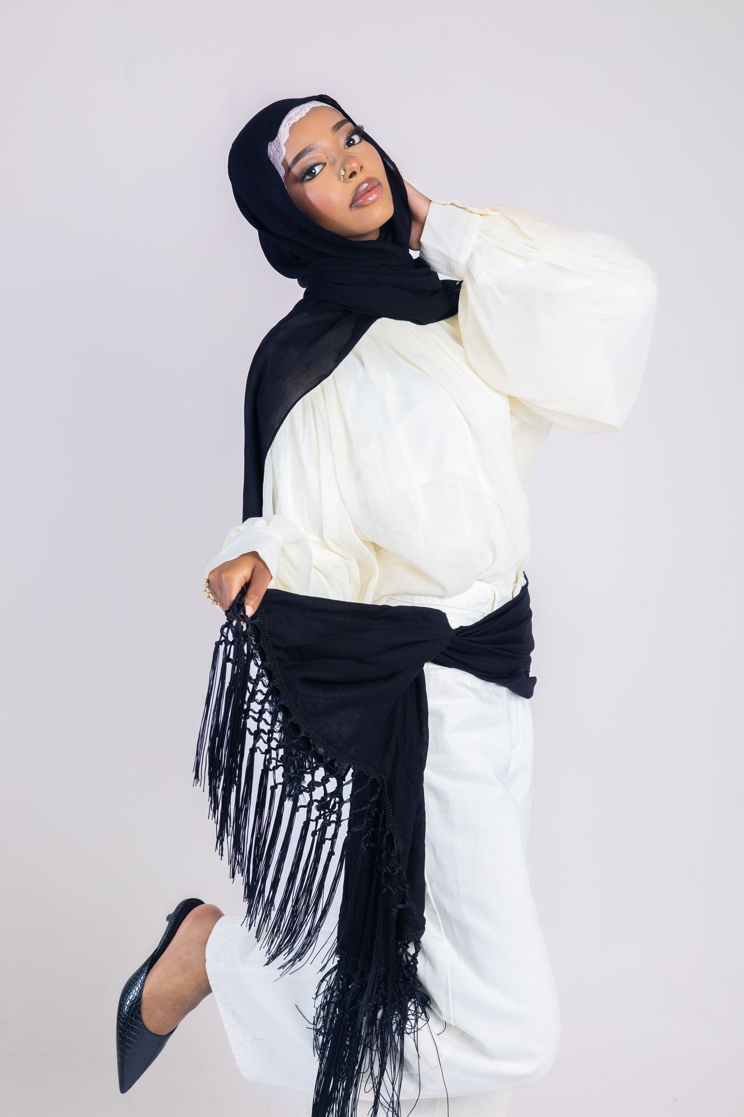 Woman in black hijab and fringed wrap poses gracefully in white outfit against light gray background.